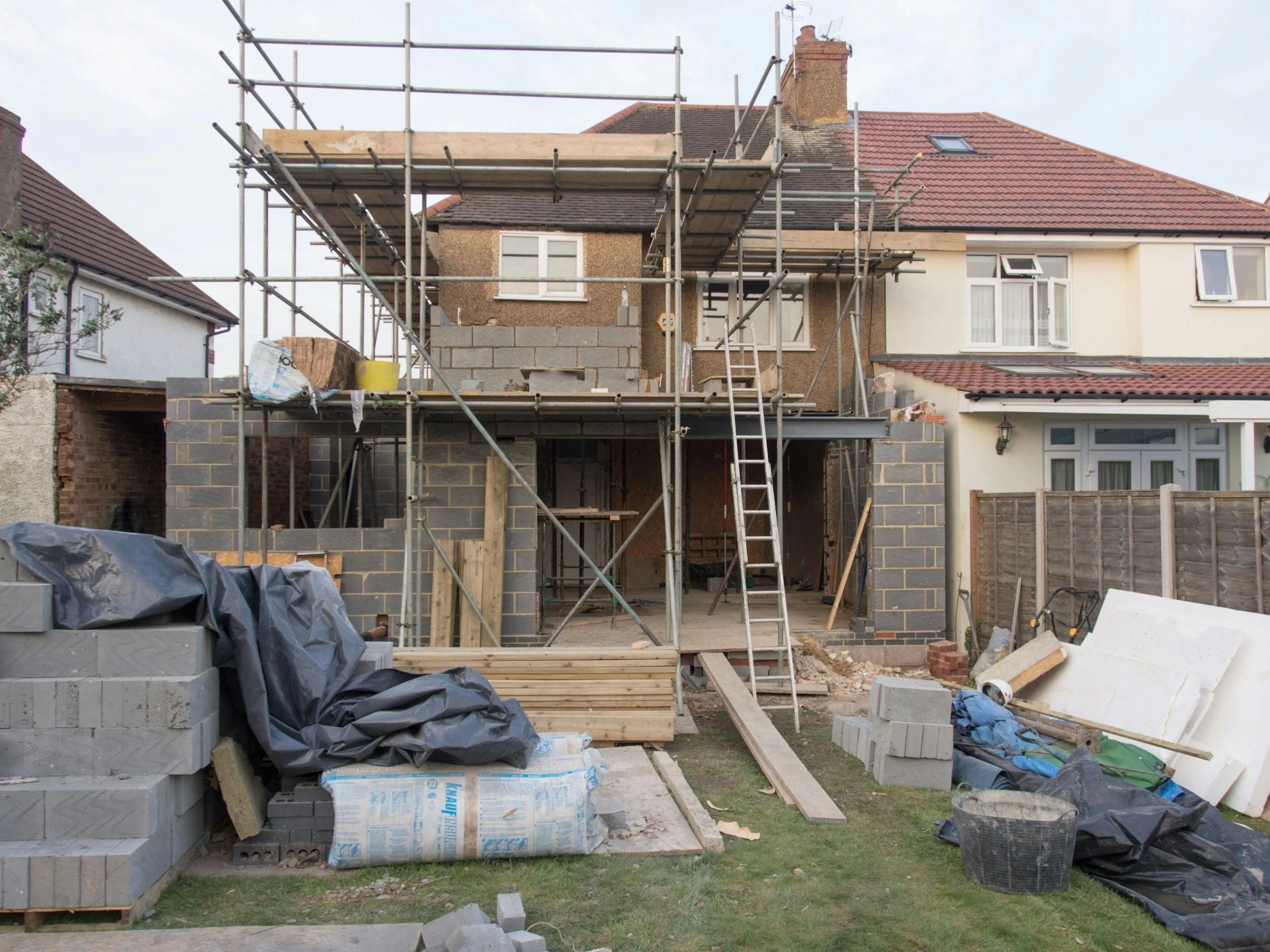 Construction site of a house with scaffolding, building materials, and equipment. The house is partially built, with bricks and cinder blocks visible, and neighboring houses are also visible.
