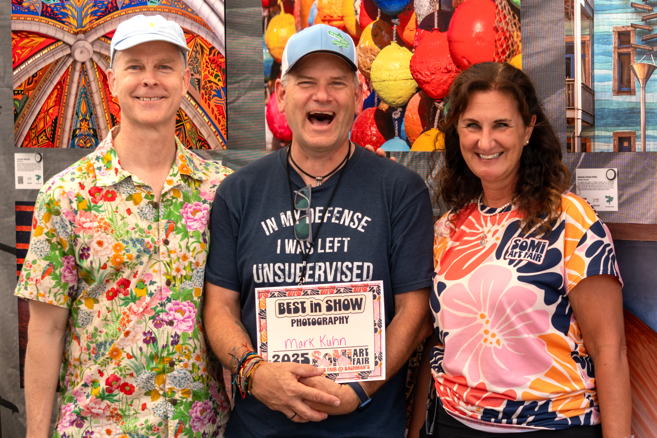 Three people at an art fair, two men and one woman, smiling and standing close together. The man in the center is holding a certificate that says 'Best in Show' for photography, with his name, Mark Kuhn. The background features colorful artwork and display signs.