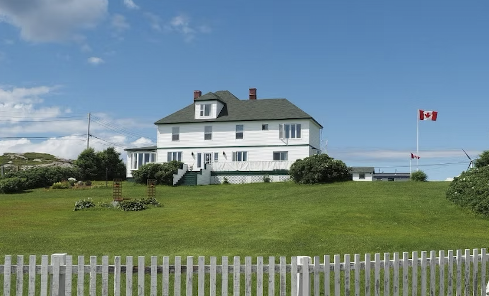 The large white Four Winds Inn sitting on the top of a hill, in front of a white picket fence.