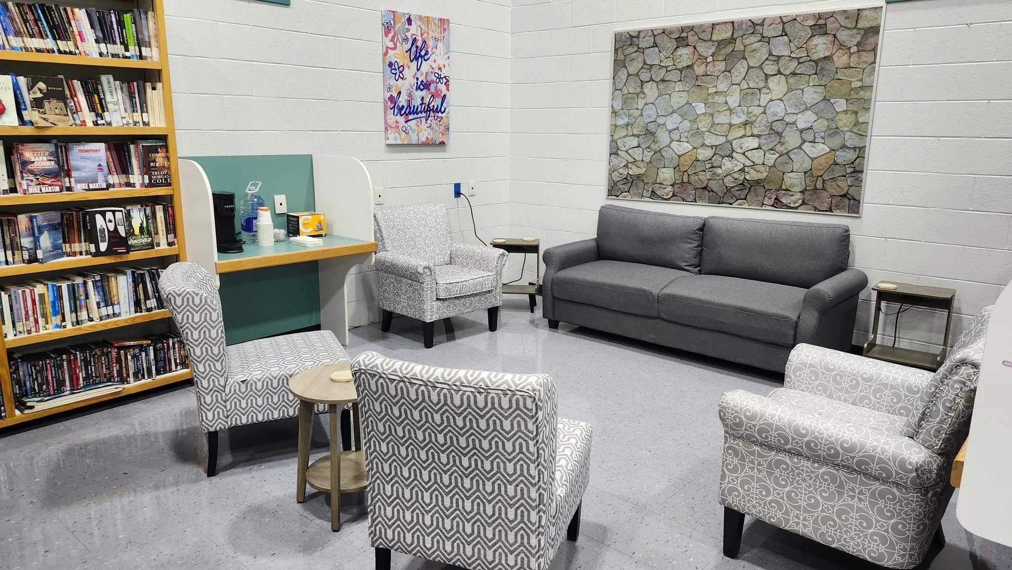 A cozy waiting room with patterned armchairs, a gray sofa, a small table, a bookshelf filled with books, a wall with artwork, and a corner with beverages and snacks.