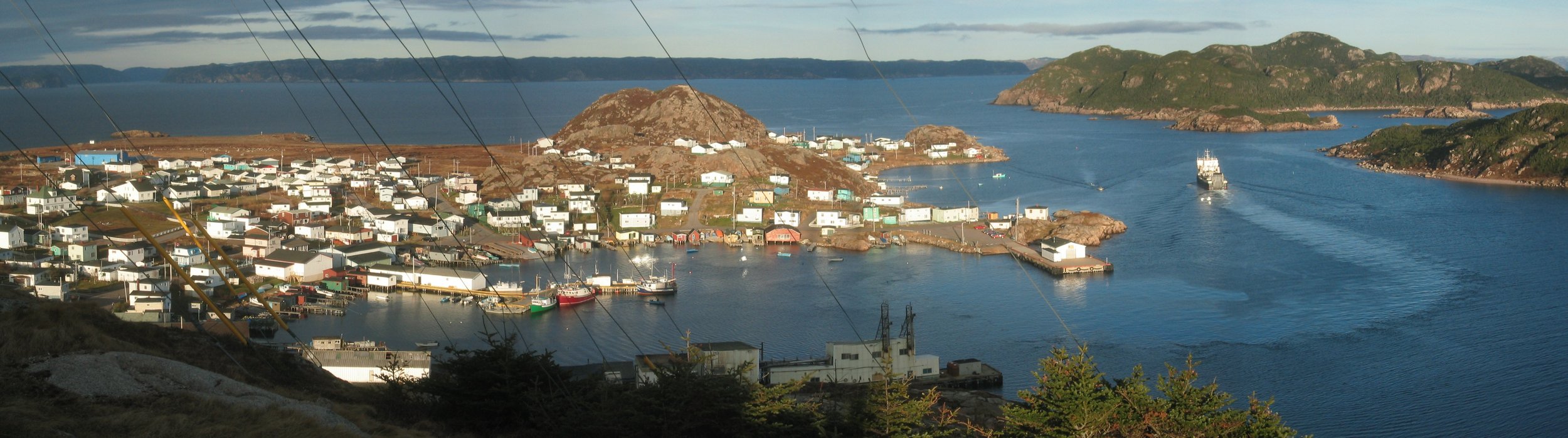 Ramea with many white houses on a hillside near the water, with boats docked in a harbor and a ship leaving the harbor, surrounded by rocky islands and hills under a blue sky.