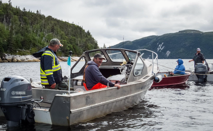 Four men on aluminum boats fishing on a lake with mountains and trees in the background.