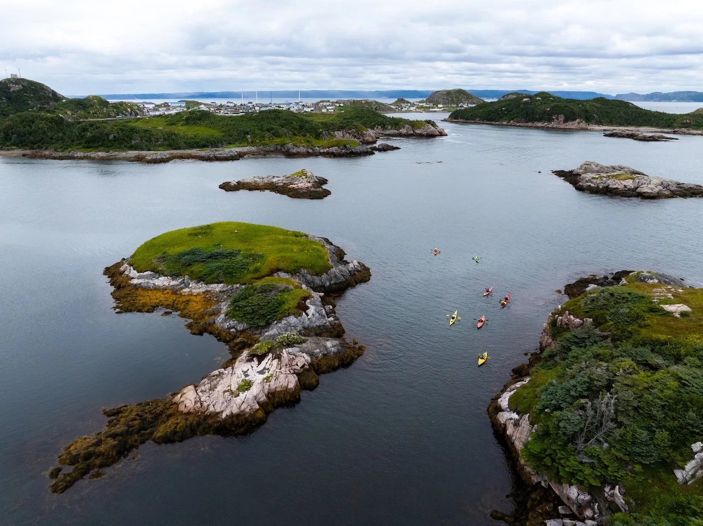 A scenic view of Ramea with small islands covered in greenery, surrounded by calm water. Several kayakers paddling in the water near the islands, with Ramea in the background under a cloudy sky.