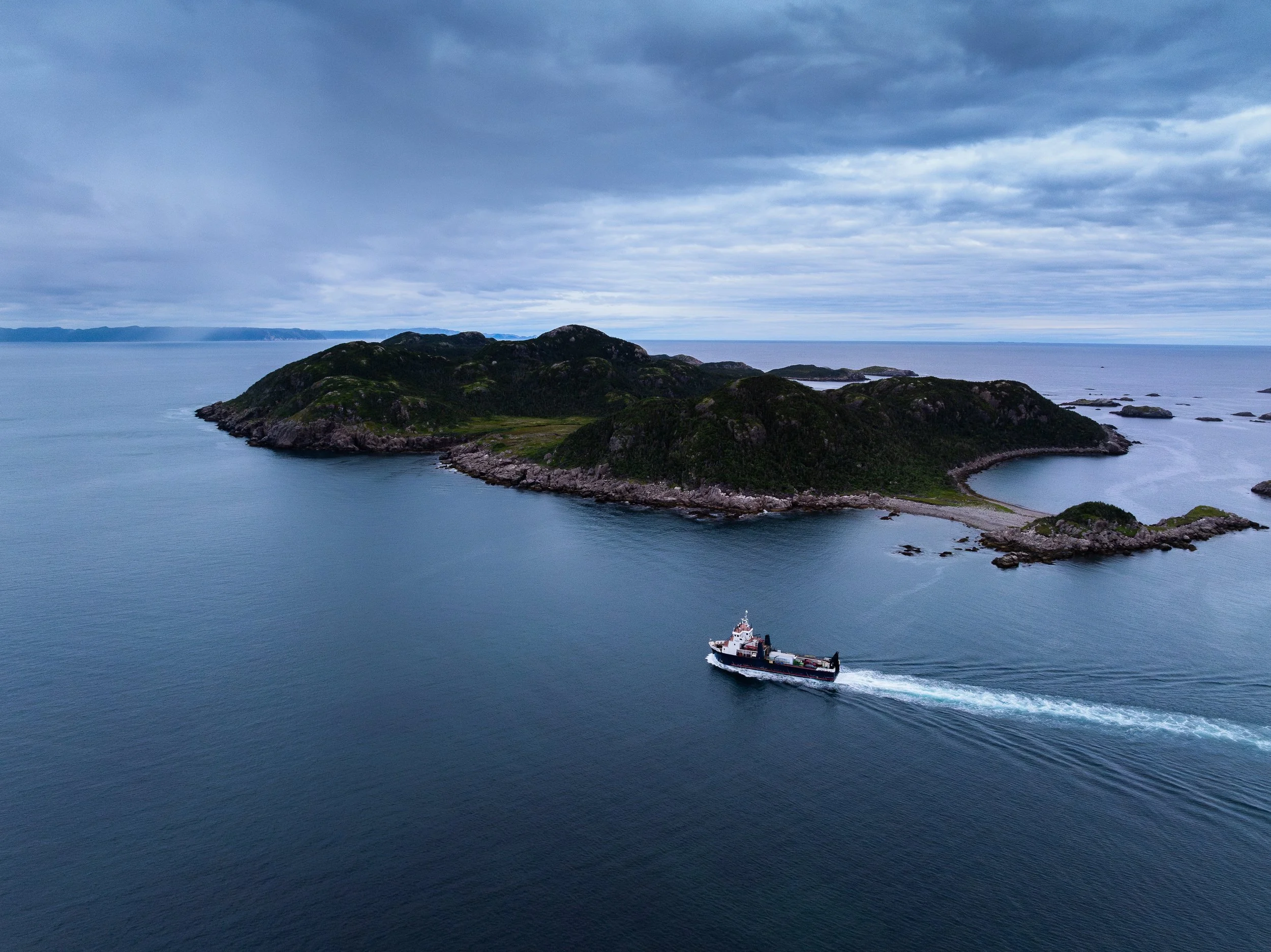 A small island off of Ramea, with green hills surrounded by water, with a ship passing nearby, under a mostly cloudy sky.