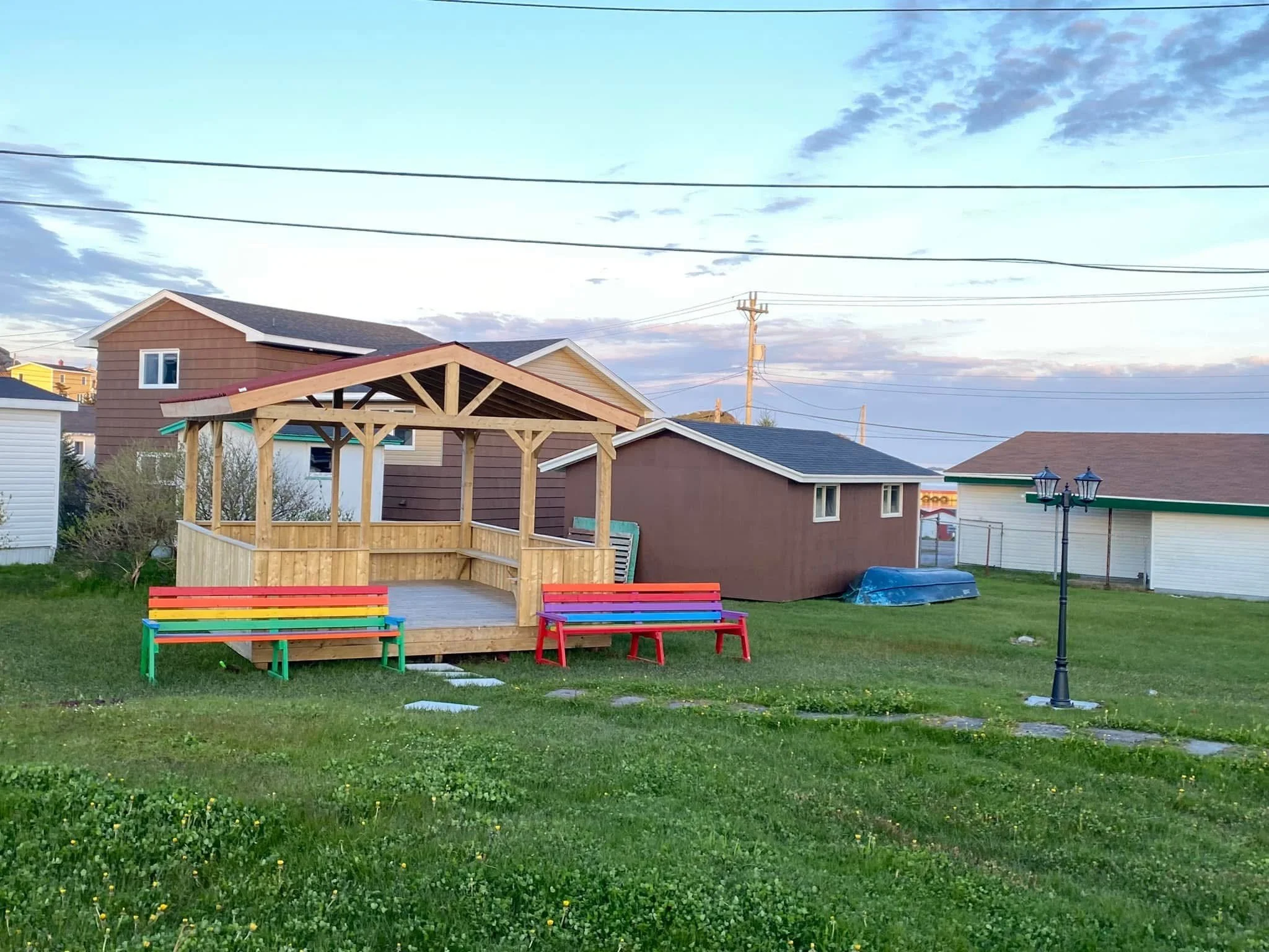 A partially built wooden gazebo with benches painted in rainbow colors is situated on a grassy lawn in a residential backyard, surrounded by houses and a lamppost.
