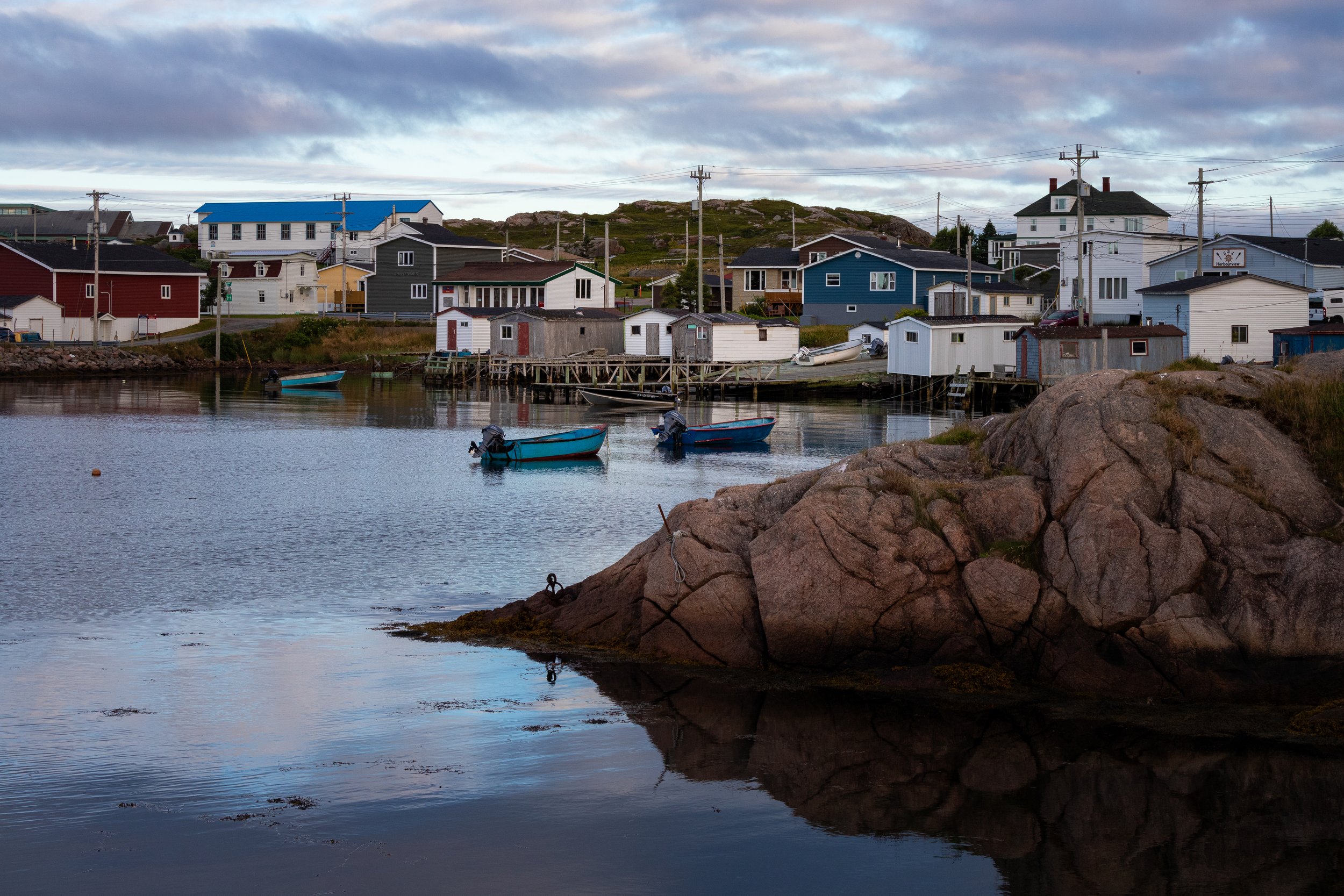A small harbor in Ramea with three boats in calm water, colorful houses on stilts along the shoreline, a rocky foreground, and a cloudy sky overhead.