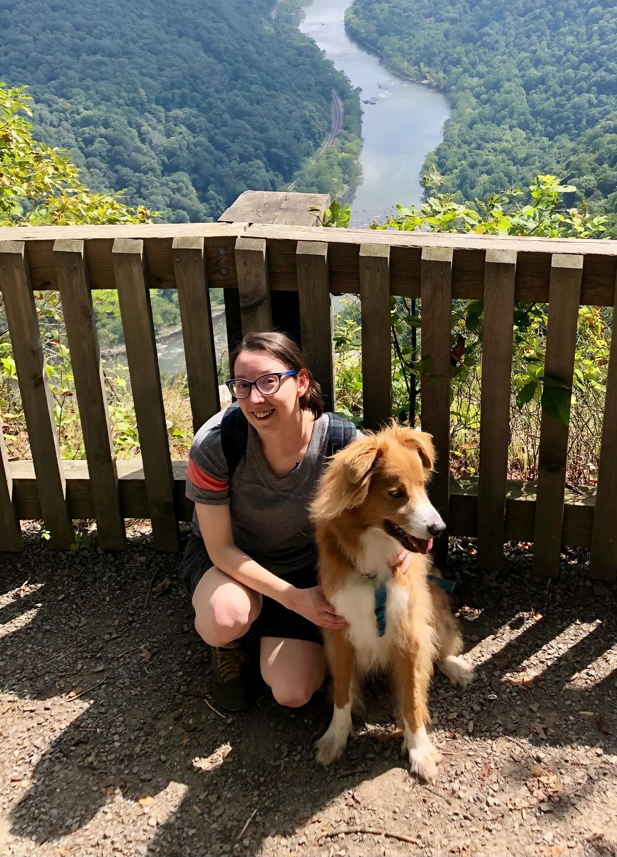 A woman wearing glasses squatting beside a golden retriever dog on a wooded overlook with a railing. In the background, there is a river winding through a lush, green valley.