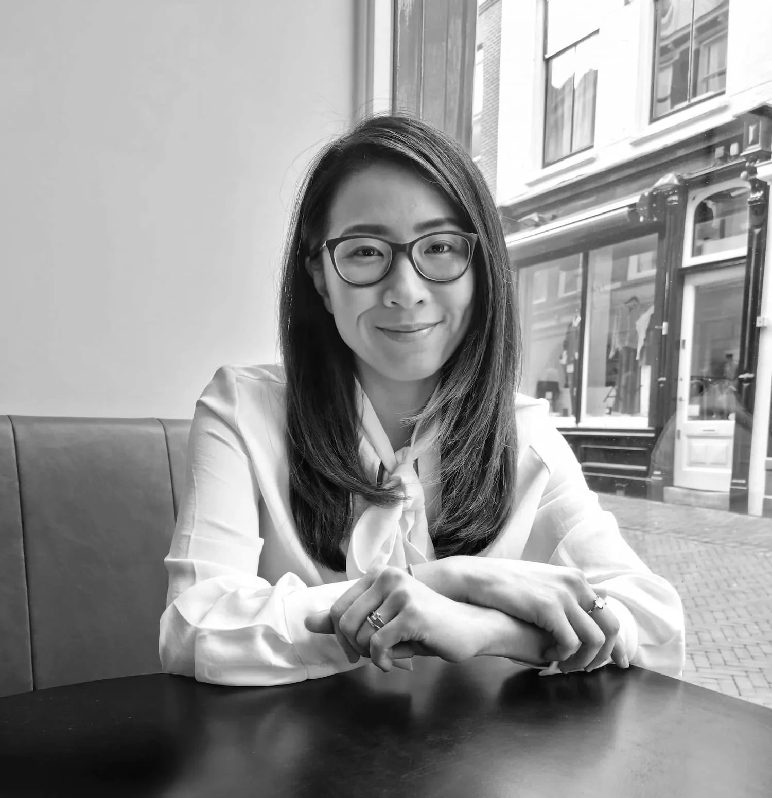 Photograph of Dr Jinnie Ooi, Chartered Clinical Psychologist, sitting at a table by a window, smiling, in black and white.