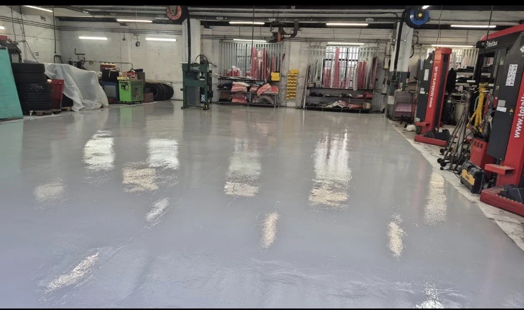 Empty industrial workshop with a clean, reflective grey epoxy floor, machinery, shelves, and equipment in the background.