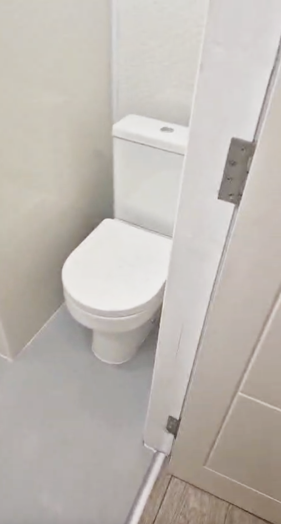 A white toilet in a small bathroom with beige walls and white flooring, partially visible inside a restroom stall with a door hinge on the right.