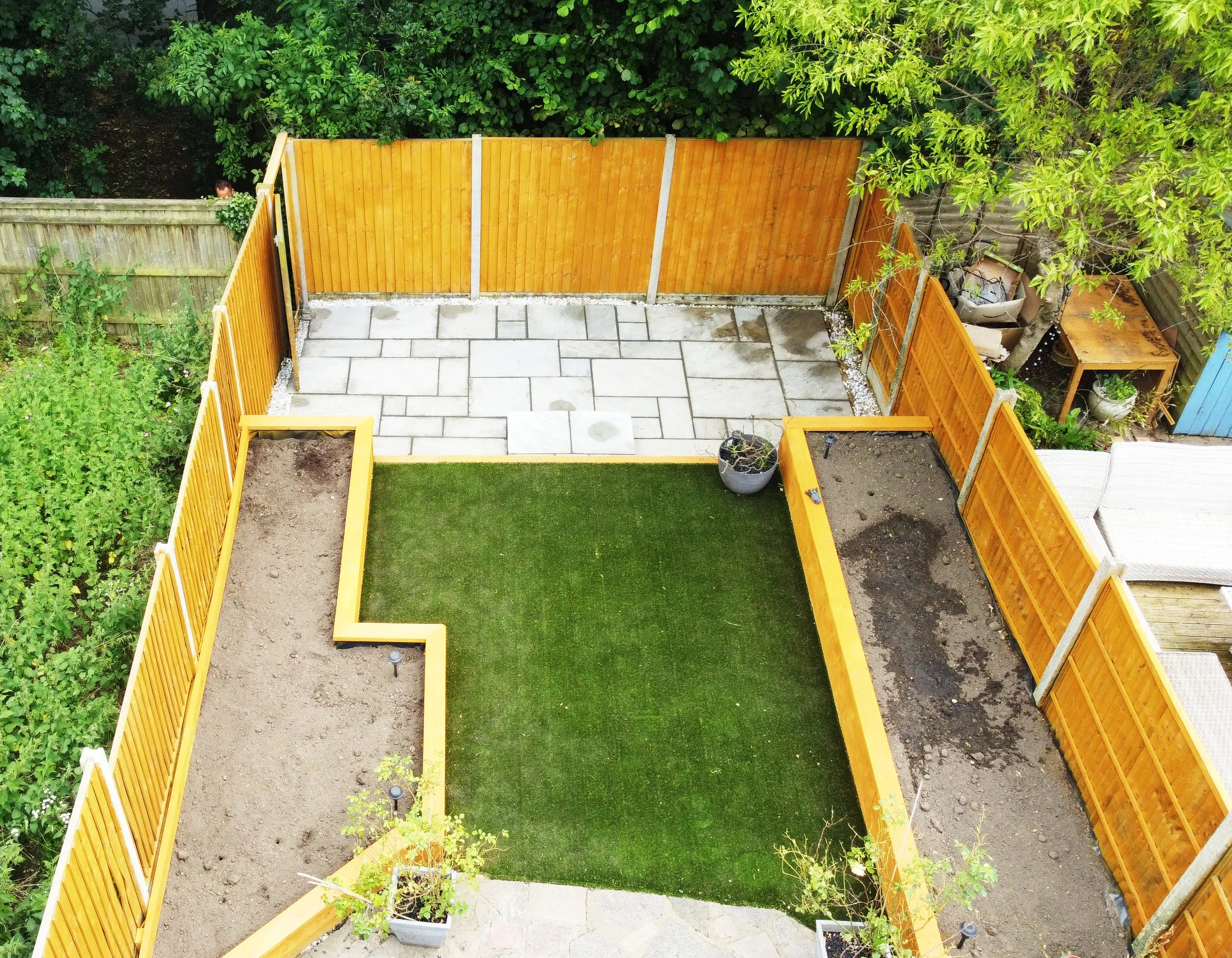 A backyard with a small patio area and a fenced lawn. The patio is paved with large, light-colored tiles, with some wet patches. Surrounding the patio are wooden fences painted yellow-orange, with a taller, natural wood fence on the left. There are t