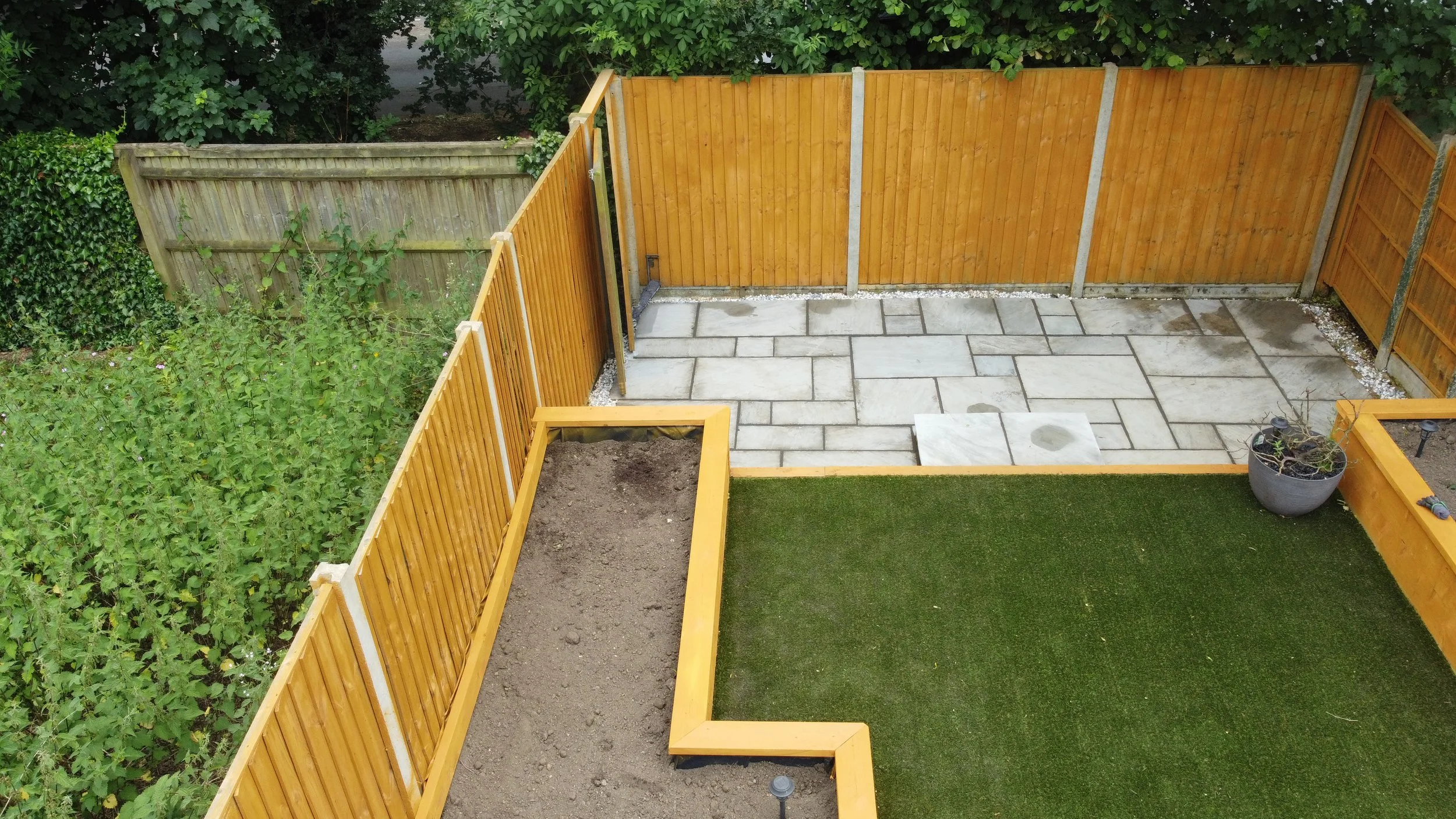A backyard with a new wooden fence, paved patio area, and a green artificial grass section, bordered by a planted flower bed and a large potted plant.