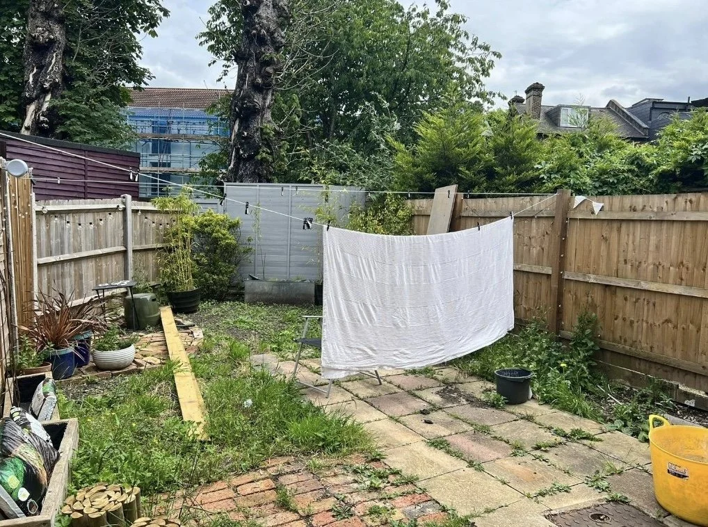 A small backyard with a clothesline, hanging white sheet, planted pots, and a stone patio surrounded by a wooden fence and trees.