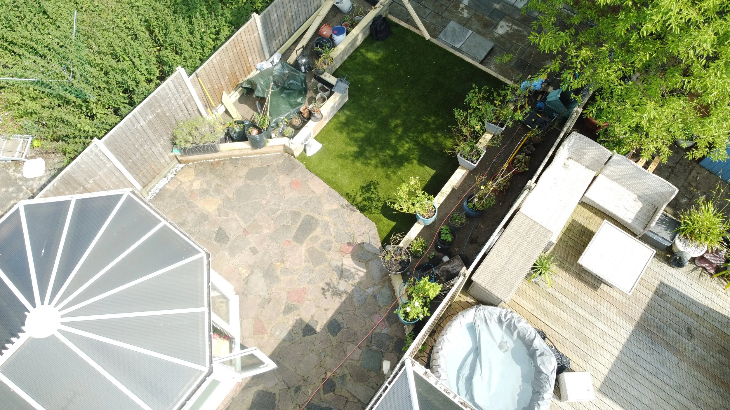 Top-down view of a backyard with a stone patio, a small grassy area, a wooden deck with outdoor furniture, potted plants, a garden shed, and a hot tub, bordered by a wooden fence and lush green trees.