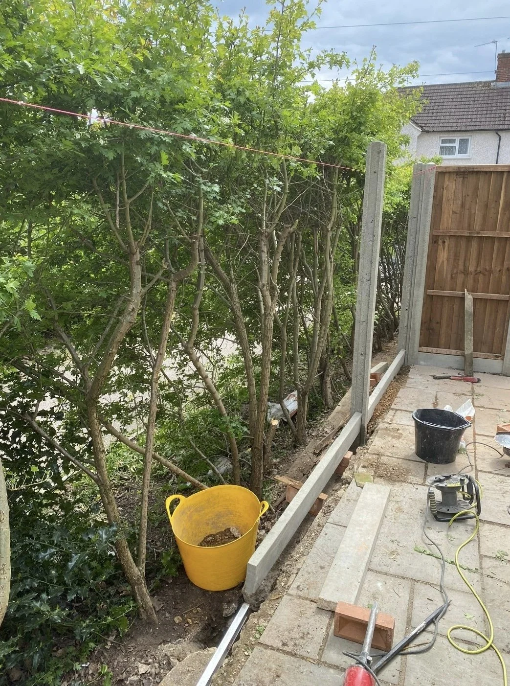 Construction site showing a partially built fence with concrete posts and wood panels, a yellow bucket, a black bucket, construction tools, and paving stones beside a row of green bushes.