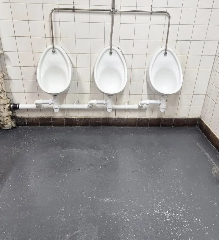 Three white urinals mounted on a tiled wall in a men's restroom with a soap dispenser on the left side and a black floor with some white paper towels.