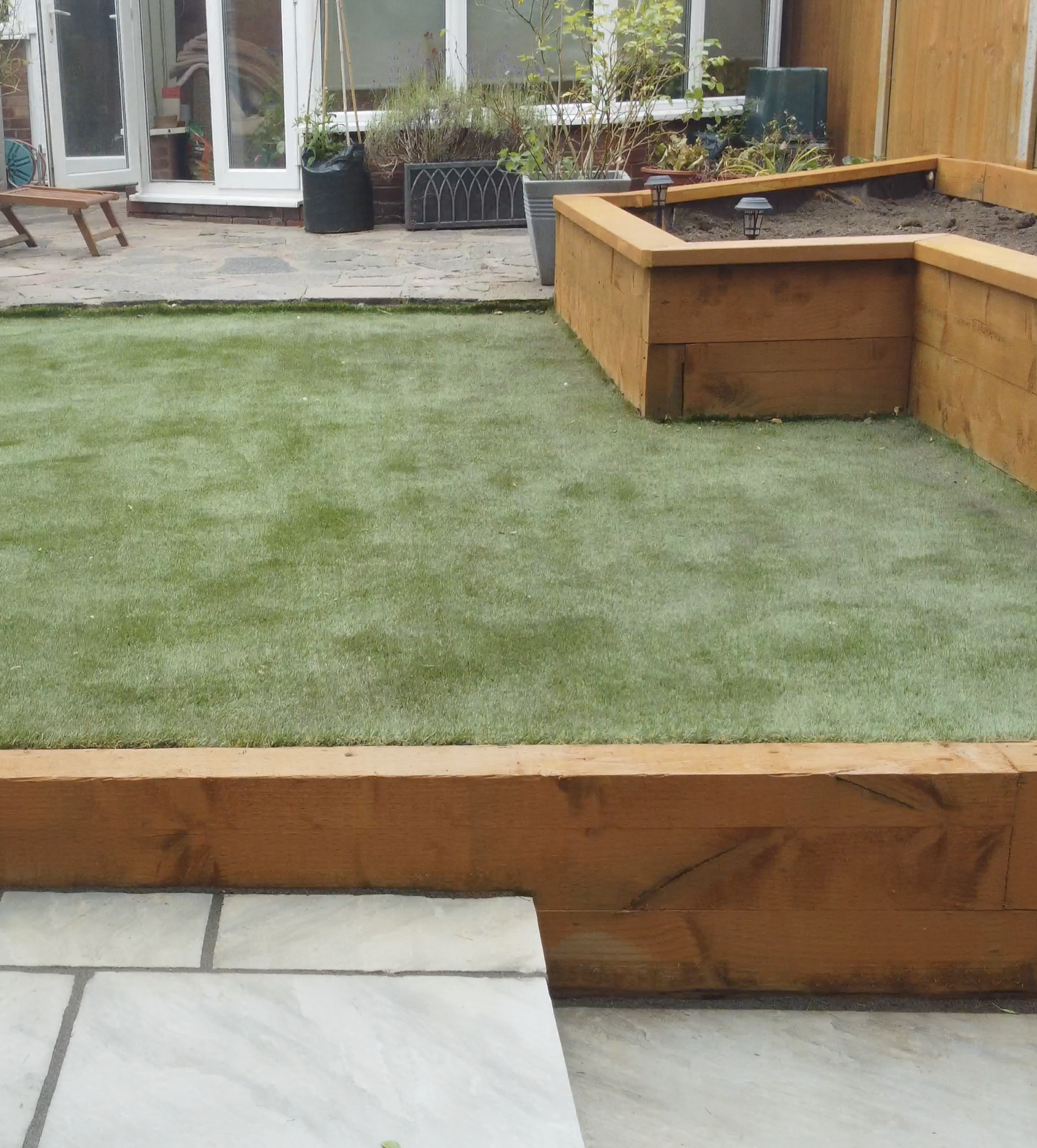 Backyard garden with artificial grass, wooden raised garden bed, potted plants, and a patio area with chairs and a glass door.
