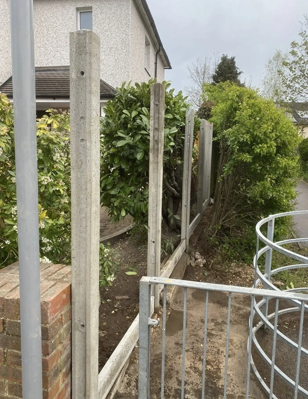 A metal fence under construction next to a garden with green bushes and a brick wall in front of a white house with a textured exterior.