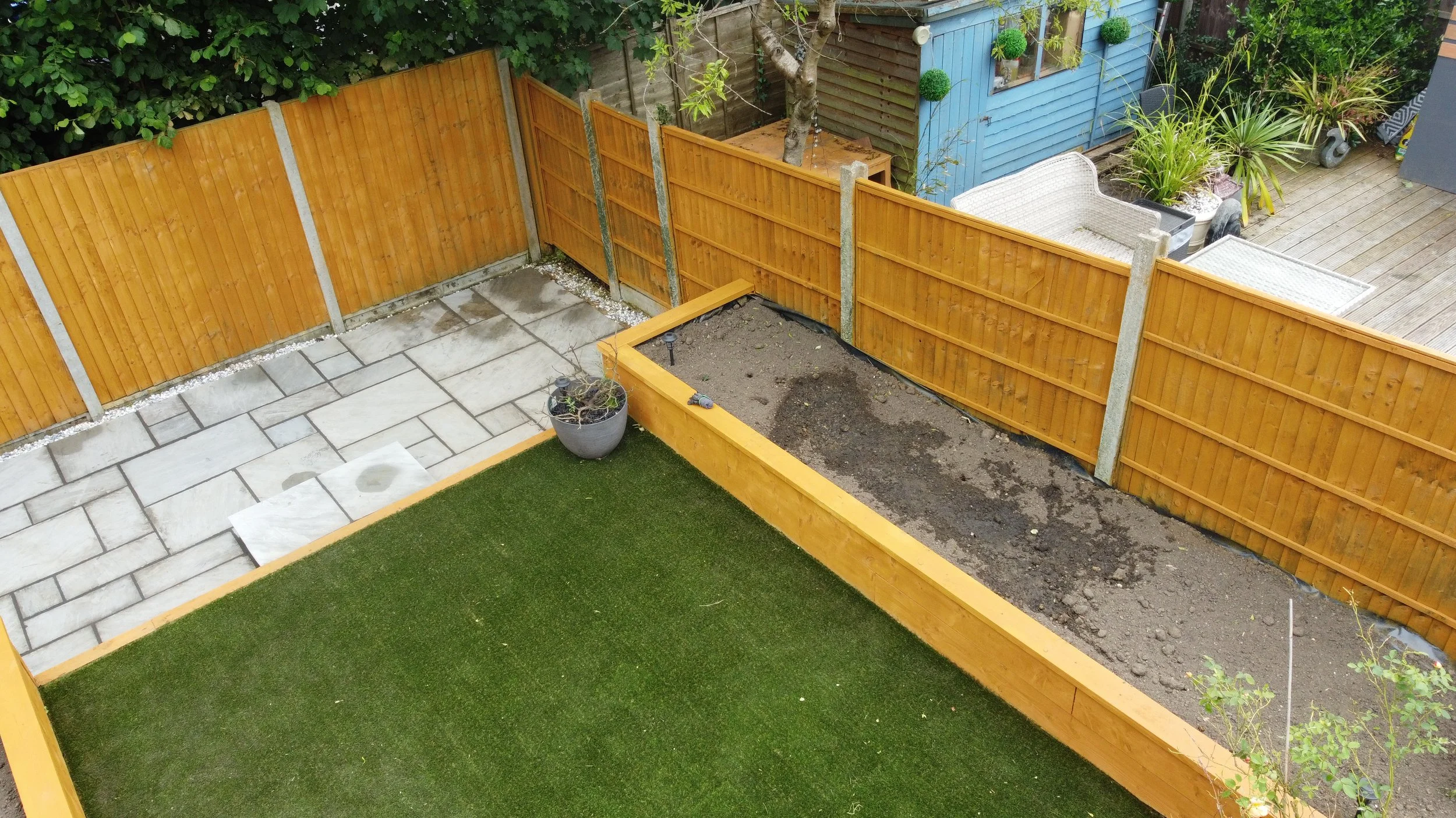 Backyard garden with a wooden privacy fence, a small raised planter bed with soil, a potted plant, an area of artificial grass, and a partly paved patio with some outdoor furniture and plants.