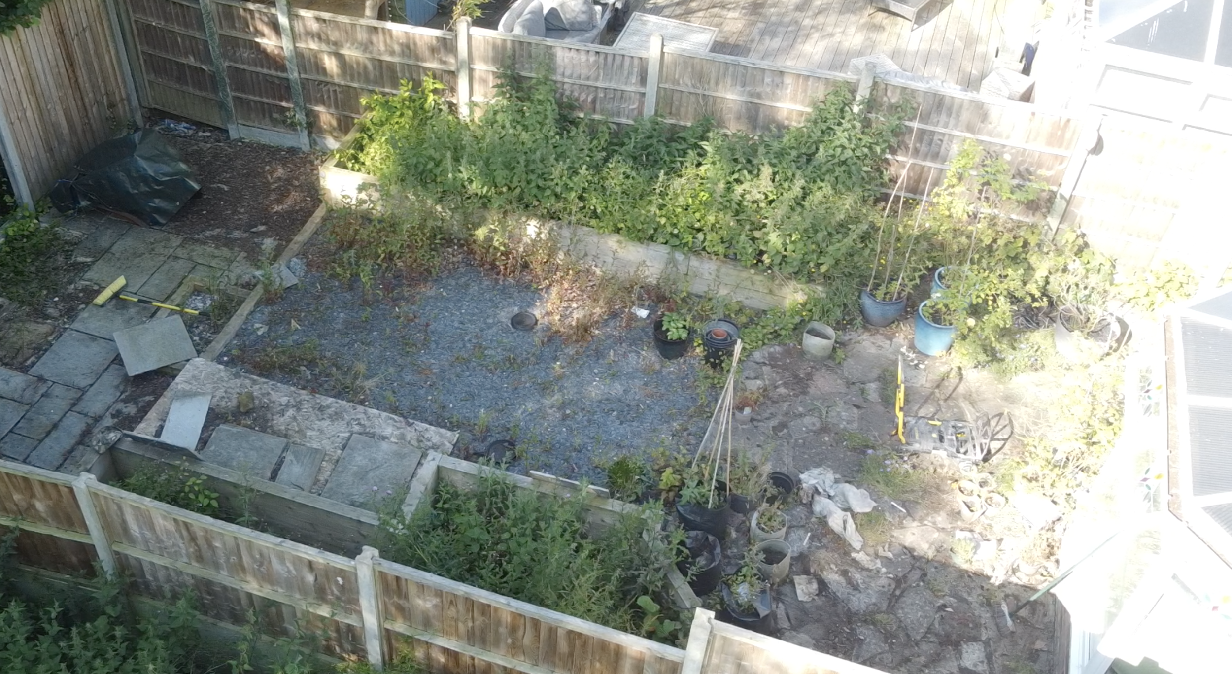 Backyard garden with plants, potted plants, wooden fence, and gravel ground. Tools and debris are scattered around.