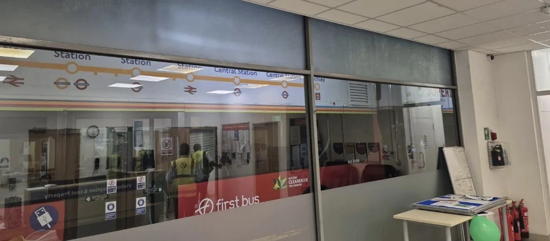 View of a train station ticketing area with glass doors, a sign for First Bus, and reflections of people inside the station.