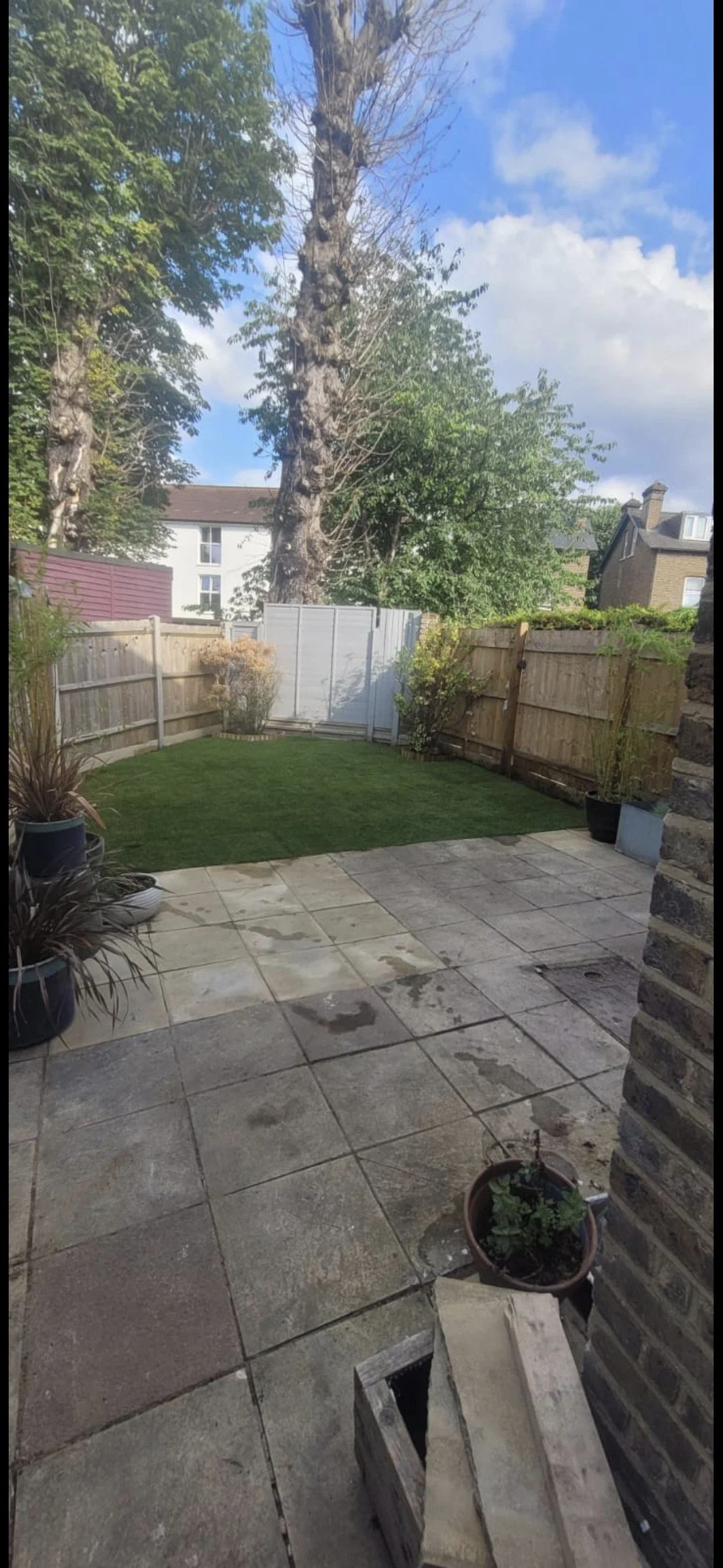 A backyard with a tiled patio area, a small patch of grass, potted plants, and a wooden fence. There is a large tree, some houses in the background, and partly cloudy sky.
