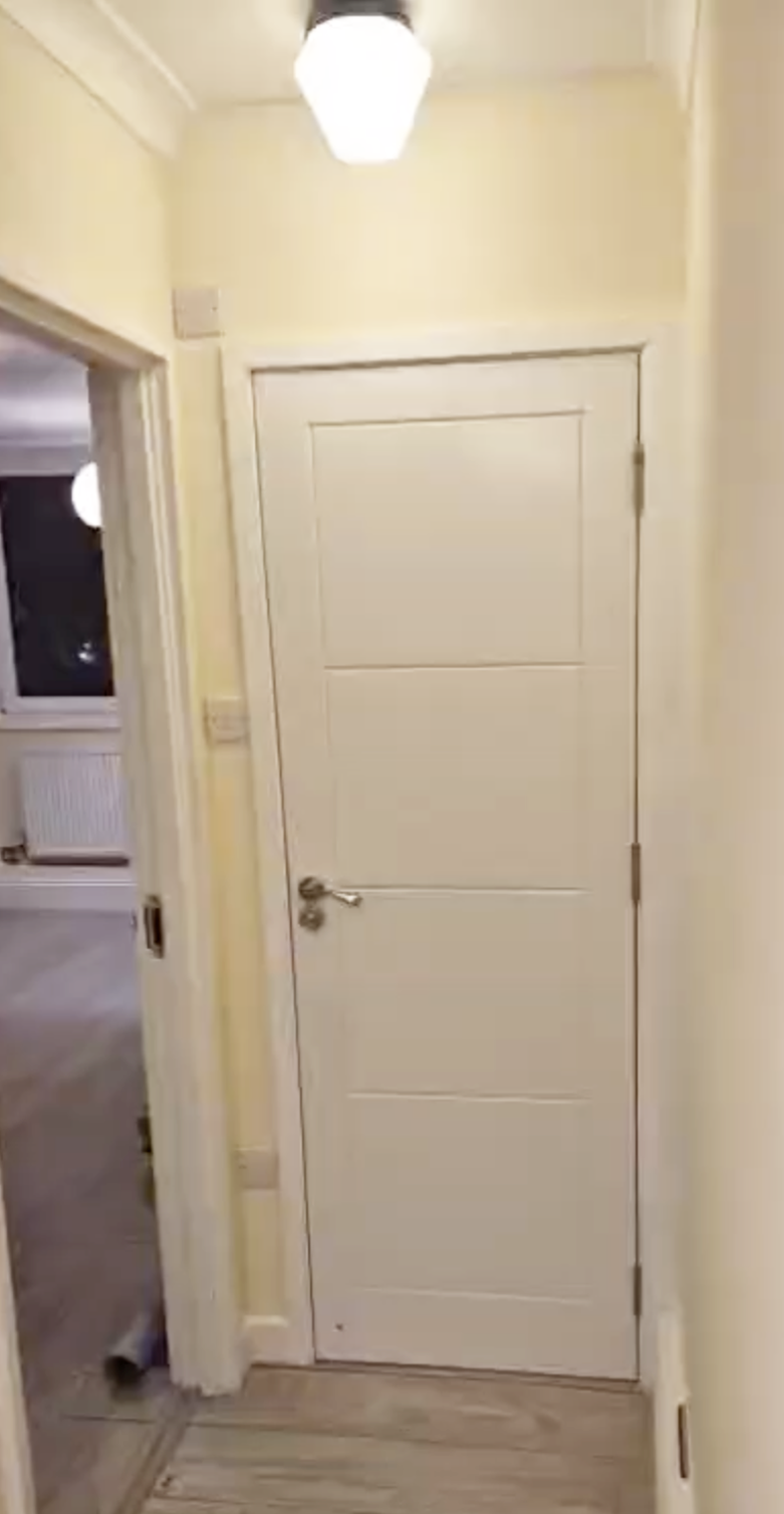 Interior hallway with a closed white door, painted yellow walls, a ceiling light, and a visible window in the adjacent room.