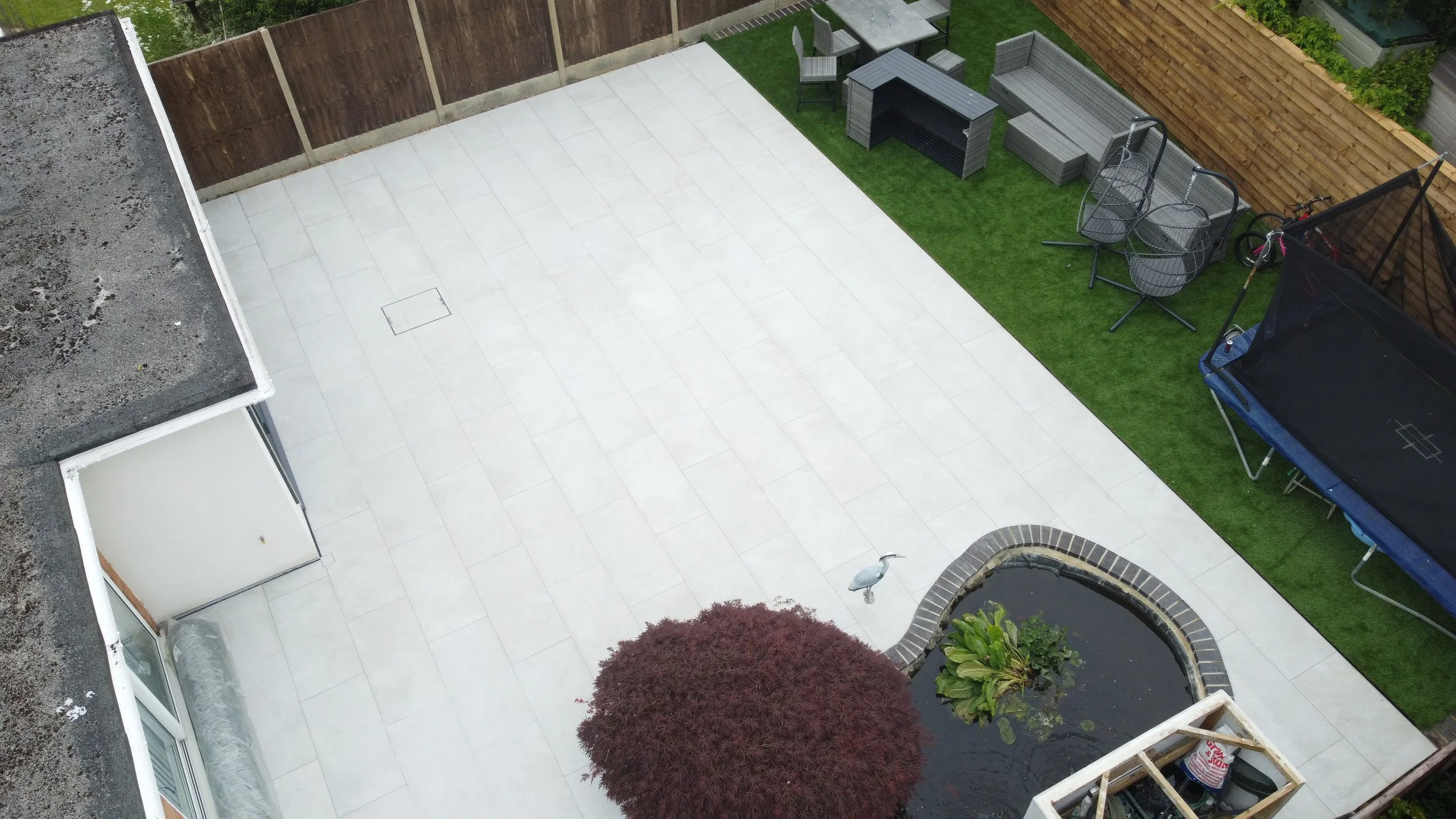 A backyard with a tiled patio area, a small pond with plants, outdoor furniture including chairs and a barbecue, a trampoline, and a wooden fence.