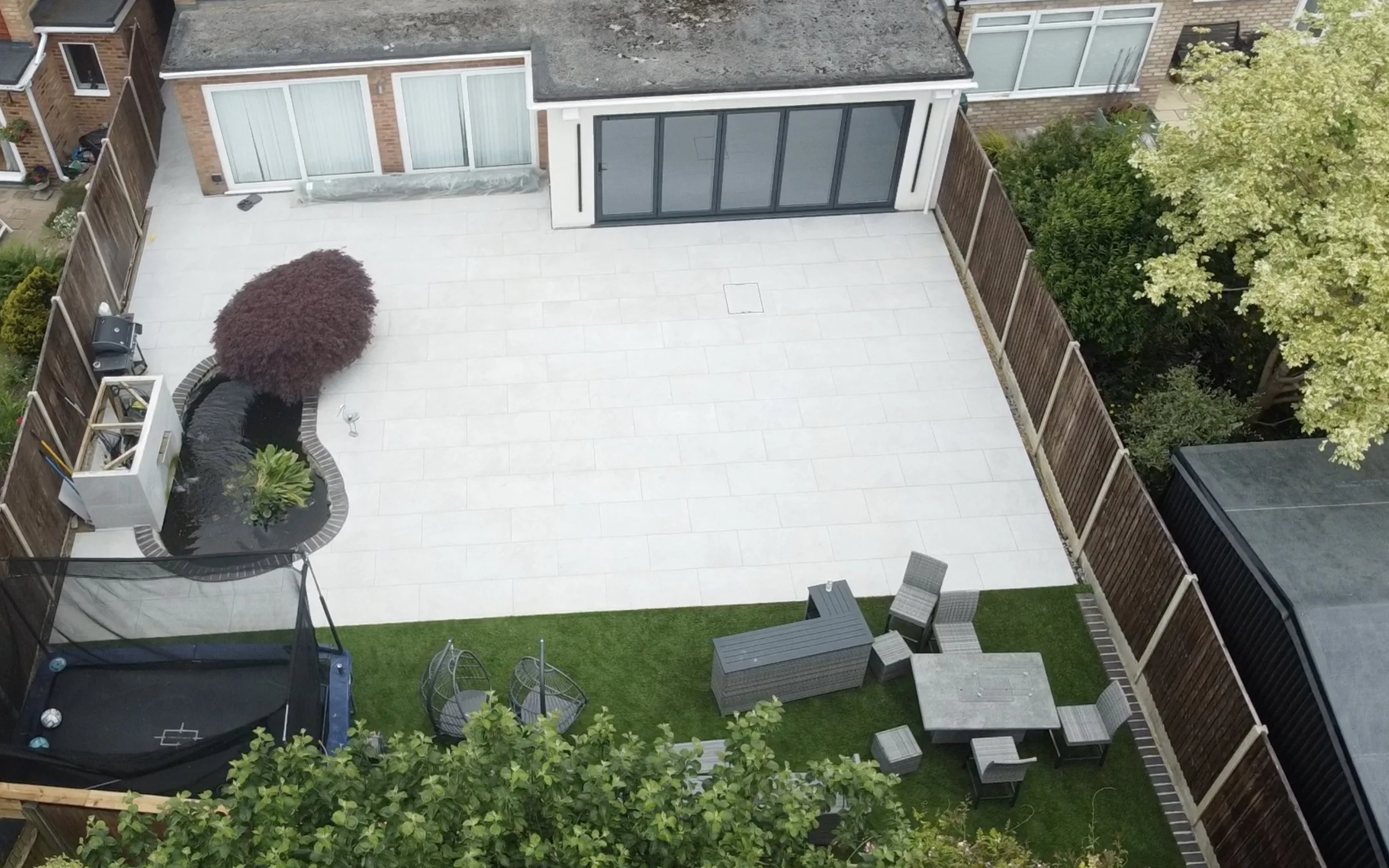A backyard with a large light-colored tiled patio, a small patch of artificial grass with outdoor seating and a trampoline, a tree with green leaves, and various plants and fences surrounding the area.