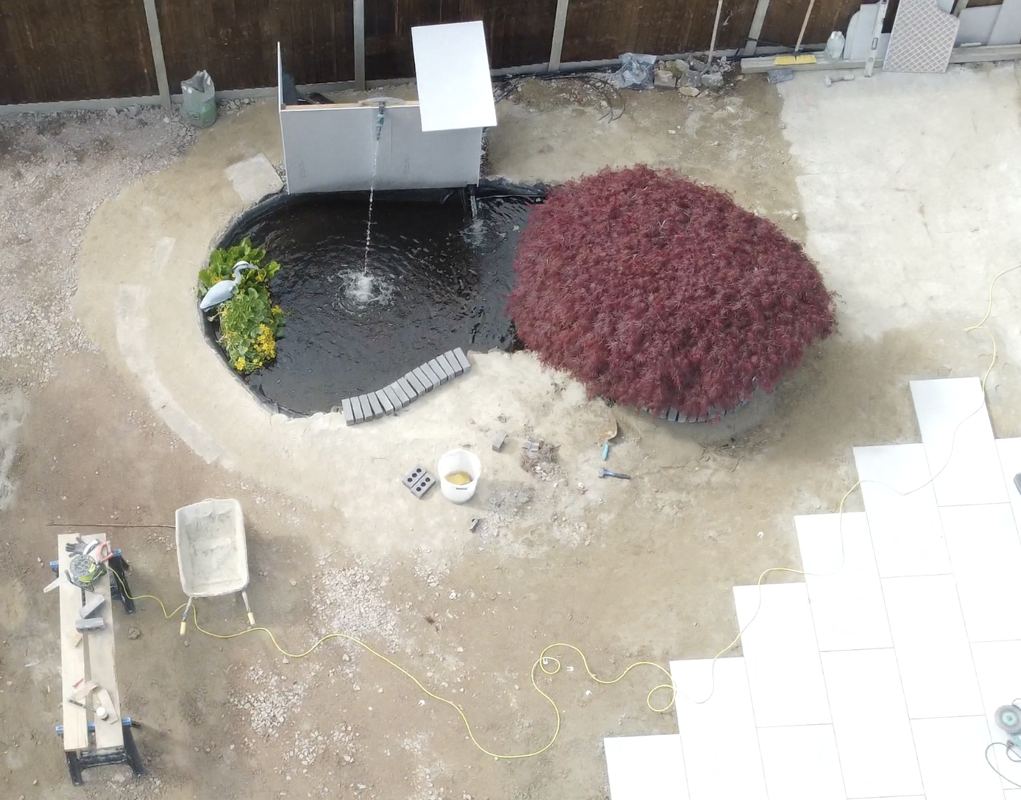 A backyard pond under construction, with a water feature, a large red bush, construction tools, and partially paved ground.