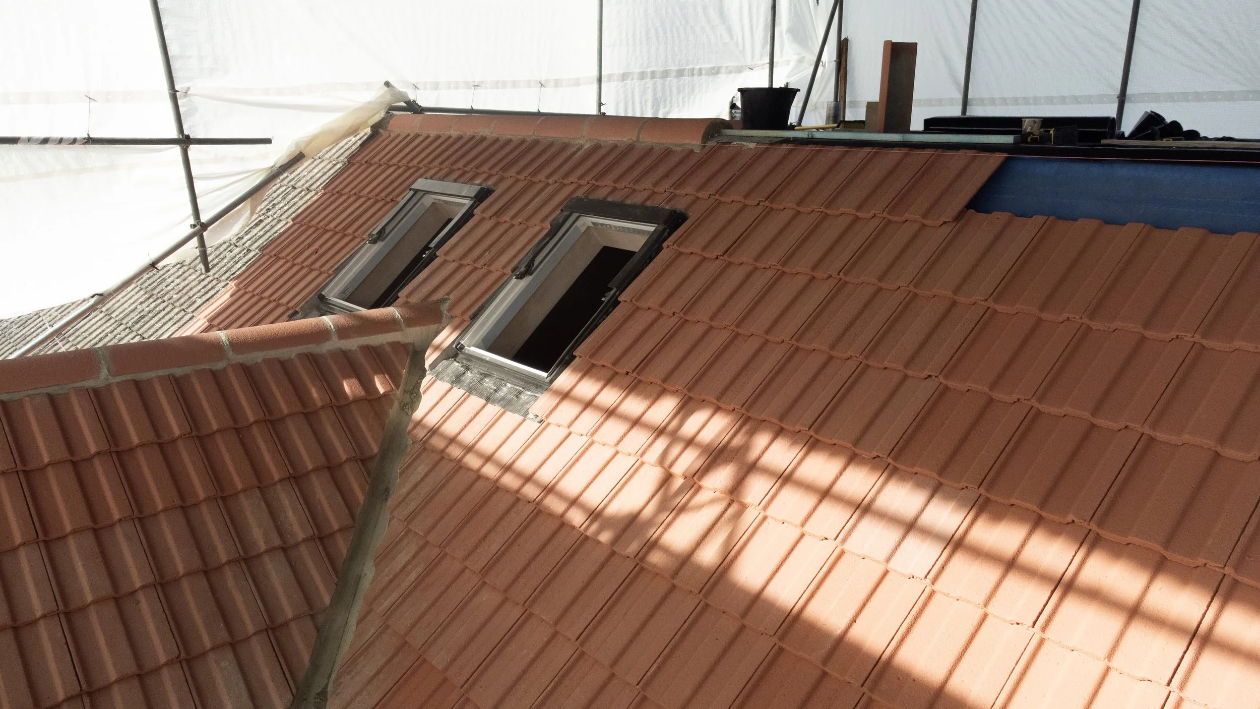 Close-up of a red tiled roof with two open skylight windows during construction, with scaffolding and building materials visible.