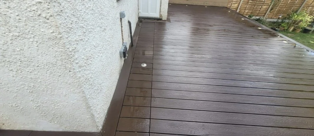 Wet wooden deck with a white door and wall on the left, and a wooden fence with potted plants on the right.