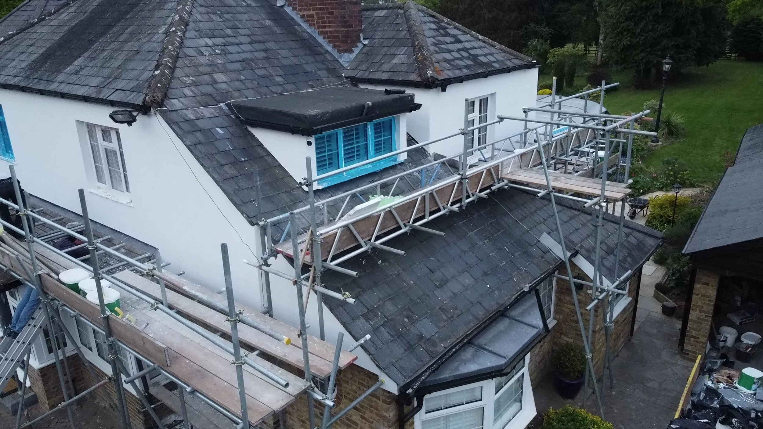 A house under renovation with scaffolding around the roof, which has dark tiles. The house has white walls and a window with blue shutters. There is construction equipment and materials on the scaffolding, and a green lawn with trees and a pathway in the background.