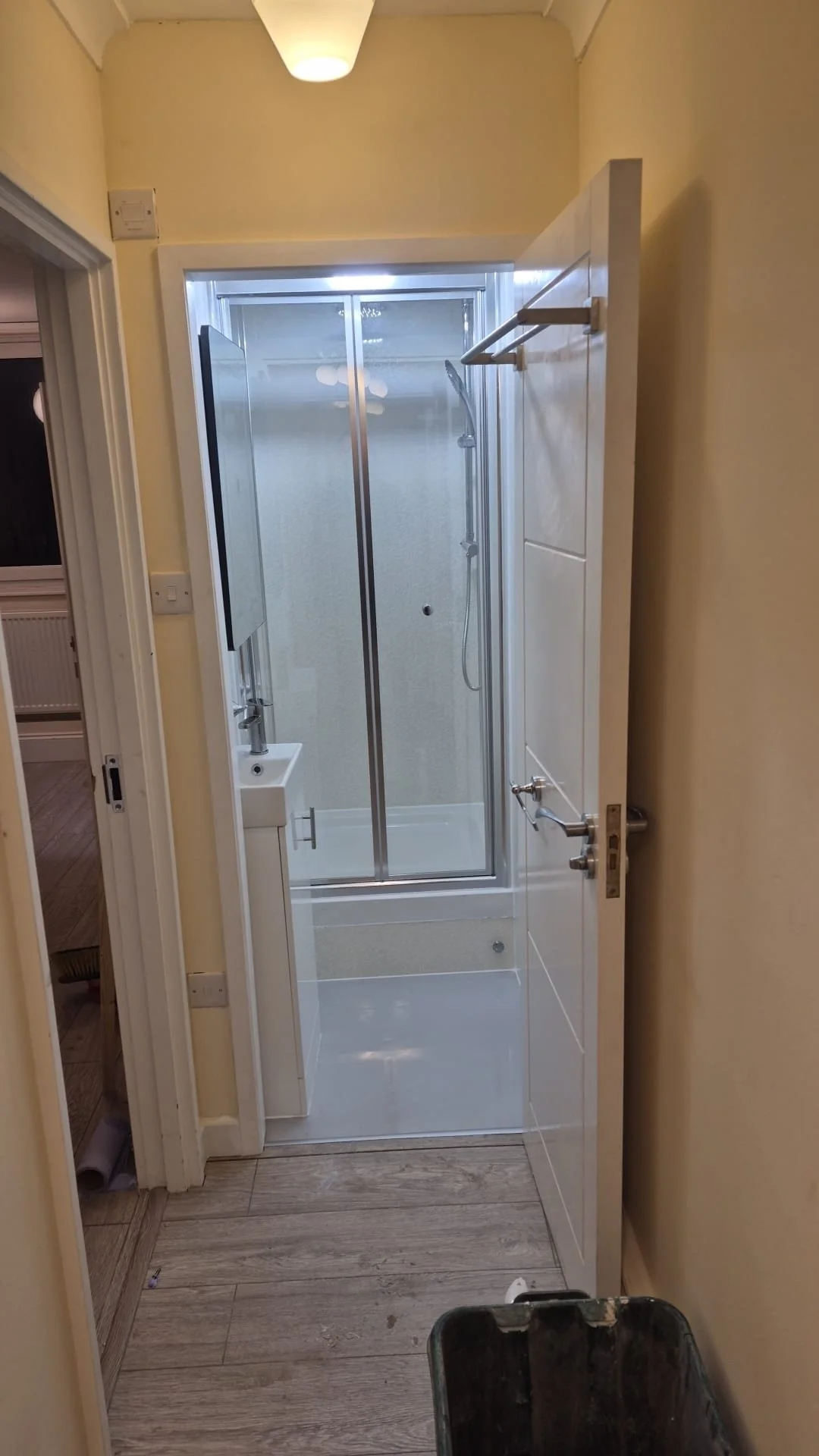 A small bathroom with a partly open door revealing a shower stall with sliding glass doors, a small sink, and a mirror. The walls are painted yellow, and the floor is wooden. There's a ceiling light fixture.