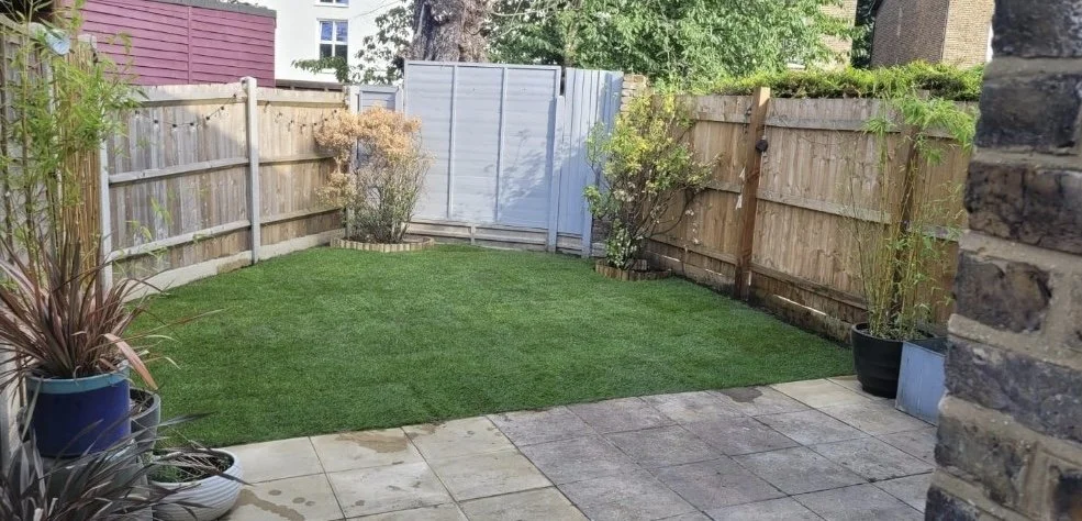 Small backyard enclosed by wooden fence with a patch of green grass, potted plants, and a stone patio area, with a shed in the background.