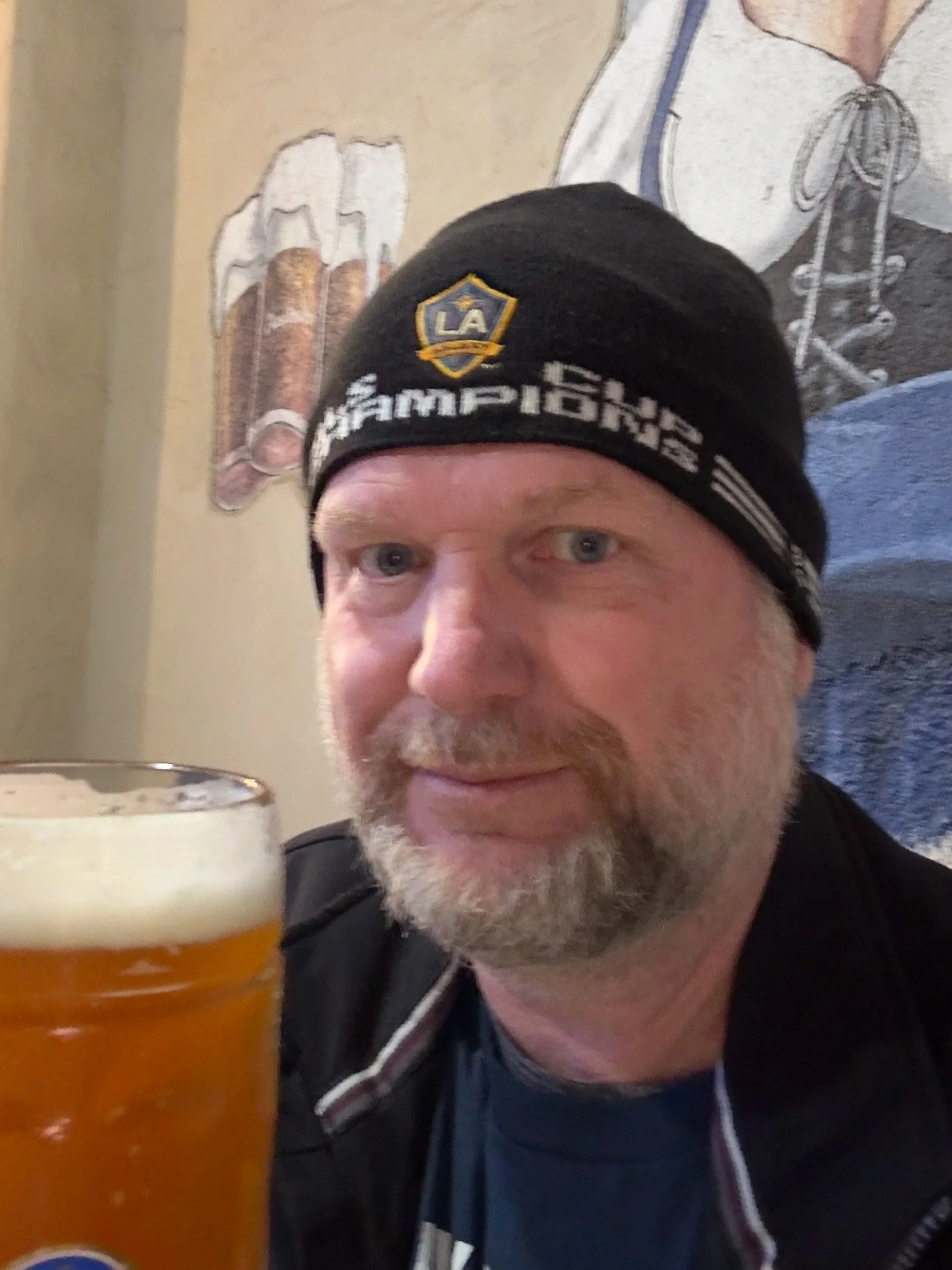 A man with a beard and blue eyes wearing a black Los Angeles Galaxy beanie sitting at a table with a glass of beer in a restaurant or pub.