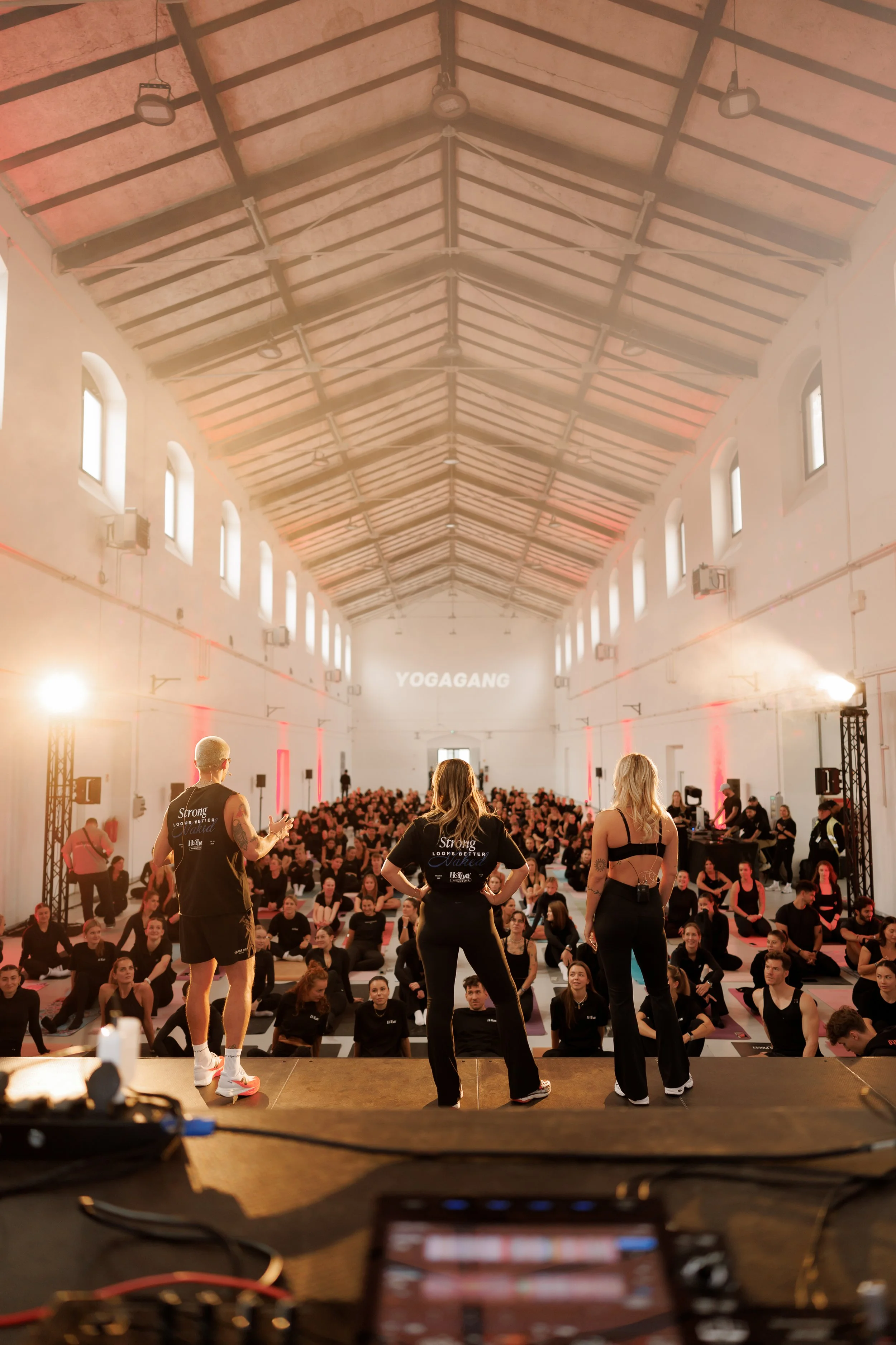 Instructor standing on stage in front of a large audience in a spacious hall with high curved ceiling, windows, and stage lighting, conducting a fitness or yoga class.