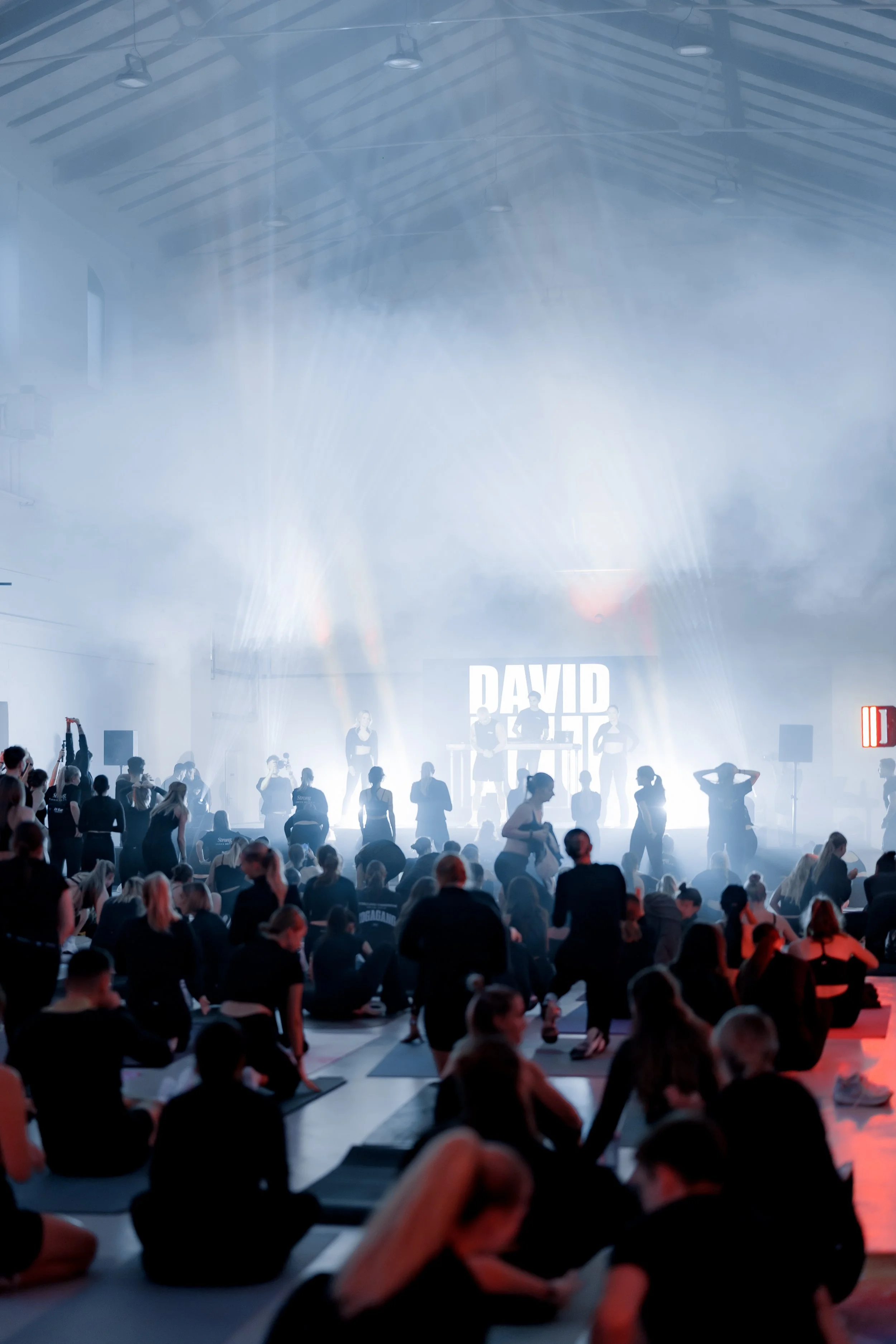 Large indoor fitness class with many participants doing exercises on yoga mats, illuminated stage at the front, large sign with the name 'DAVID' visible, bright lighting, high ceiling with exposed beams.