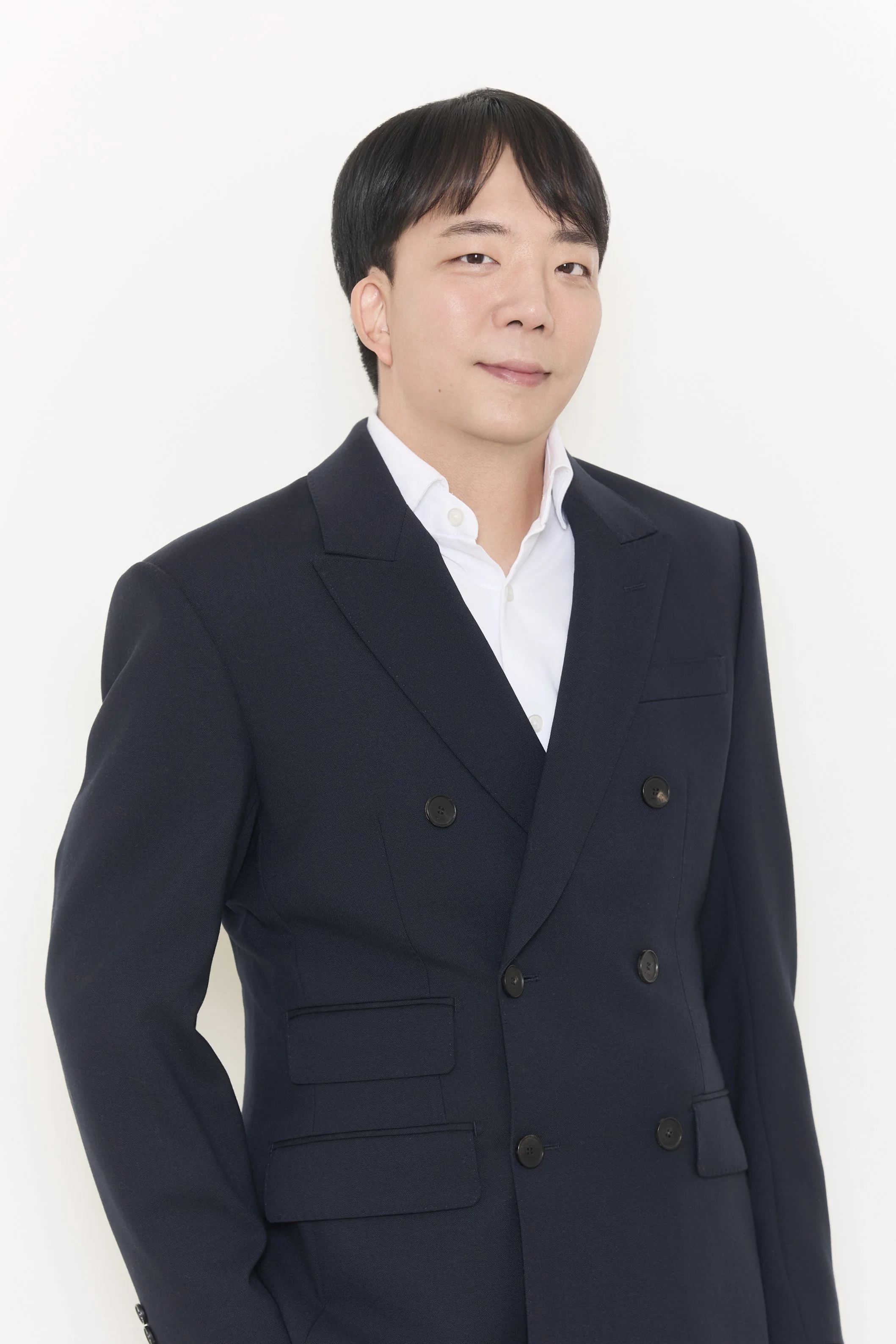A young Asian man with short dark hair, wearing a navy double-breasted blazer and a white dress shirt, standing against a plain white background.