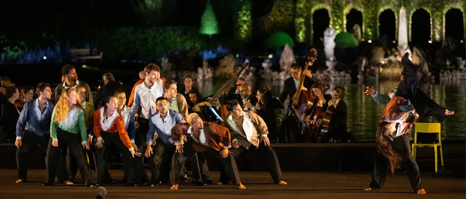 Group of performers on stage with a dance scene, including a woman performing acrobatics over a chair, and live orchestra in the background, illuminated by colorful night lighting.