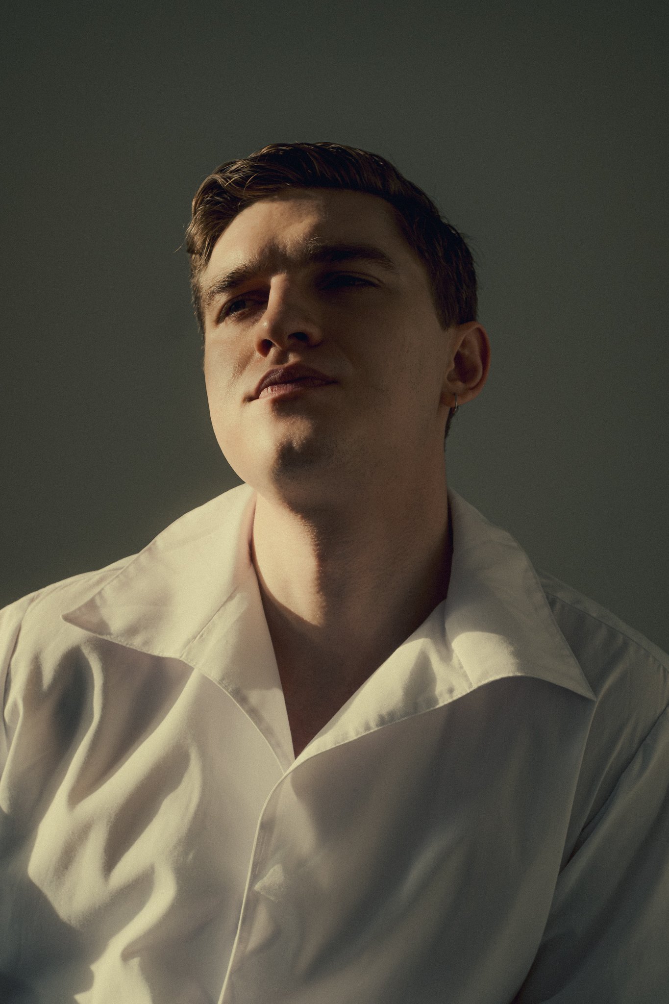Portrait of a young man with dark hair, wearing a white shirt, looking confidently upward with soft lighting.