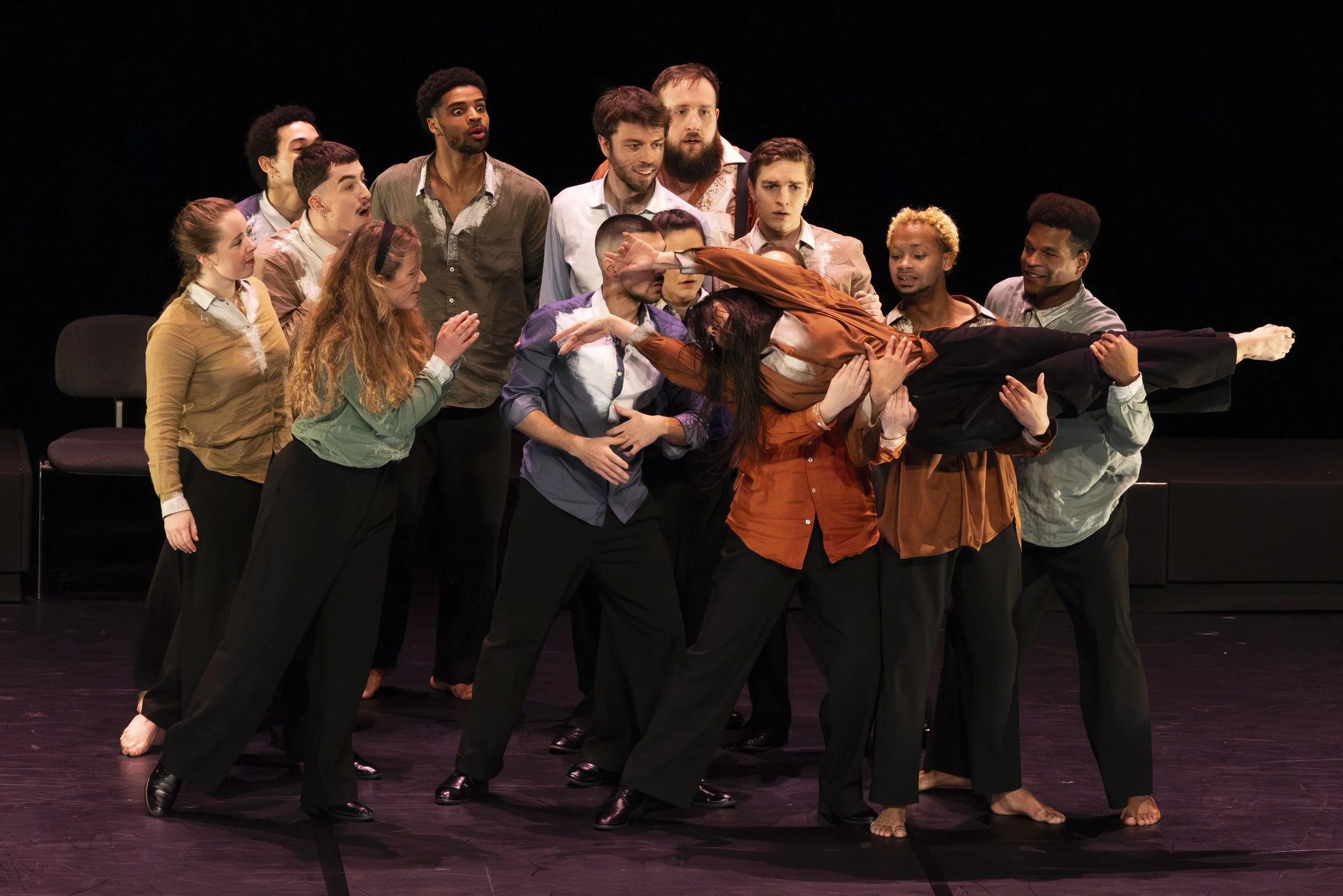 A diverse group of actors on stage performing a dramatic scene, with one person being carried horizontally by others, against a black background.