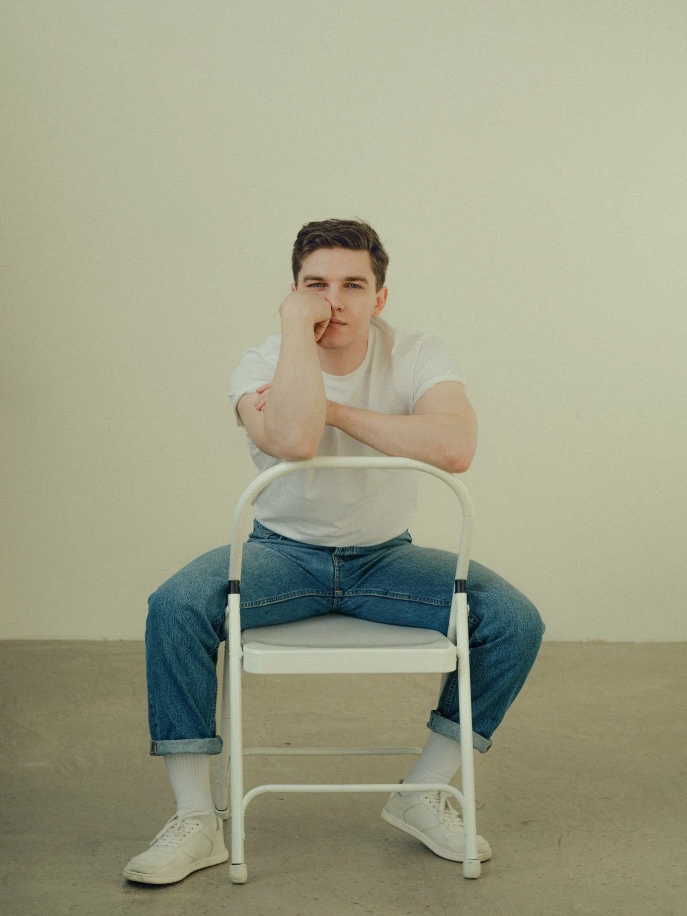 A young man in a white t-shirt and jeans sitting on a white chair against a plain light-colored wall. He is resting one arm on the chair's back and has his other hand against his face, with a thoughtful expression.