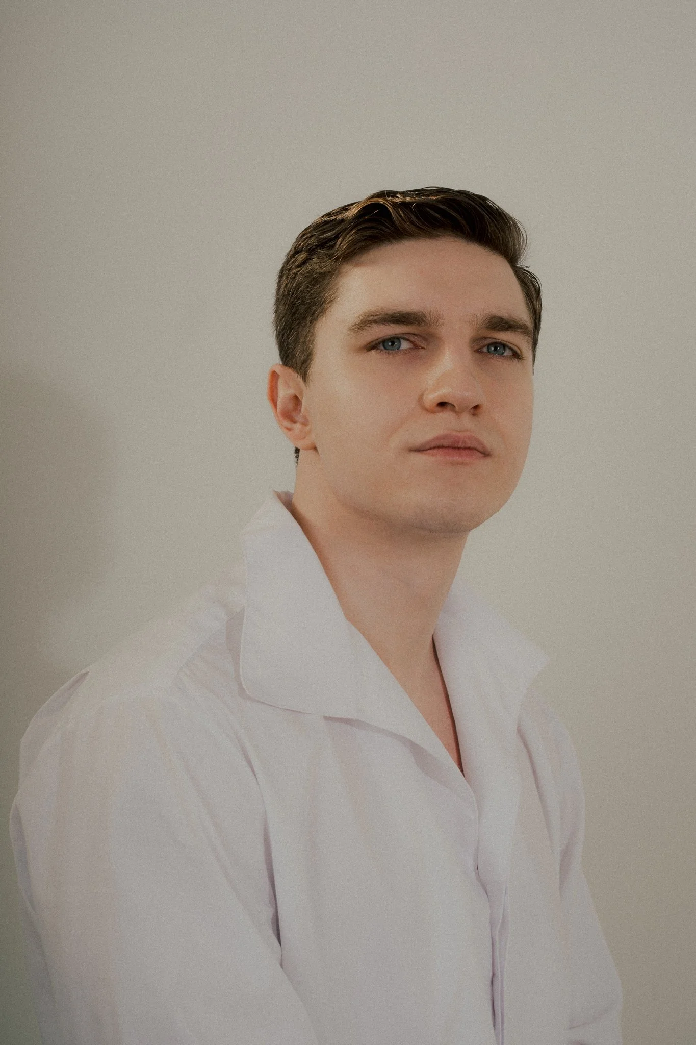 Young man with dark hair and blue eyes wearing a white shirt against a plain light background.