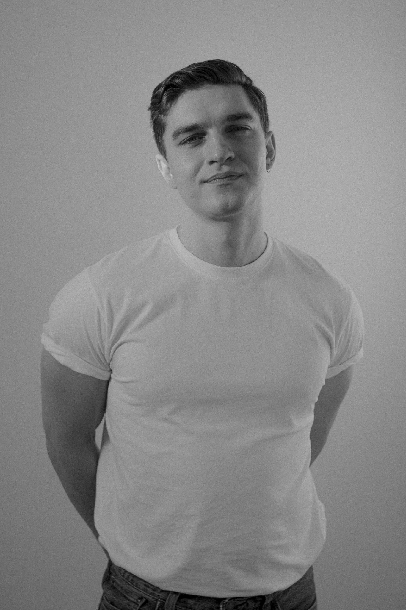 A young man with short, slicked-back hair and a slight smile, wearing a plain white T-shirt and jeans, standing against a plain wall.