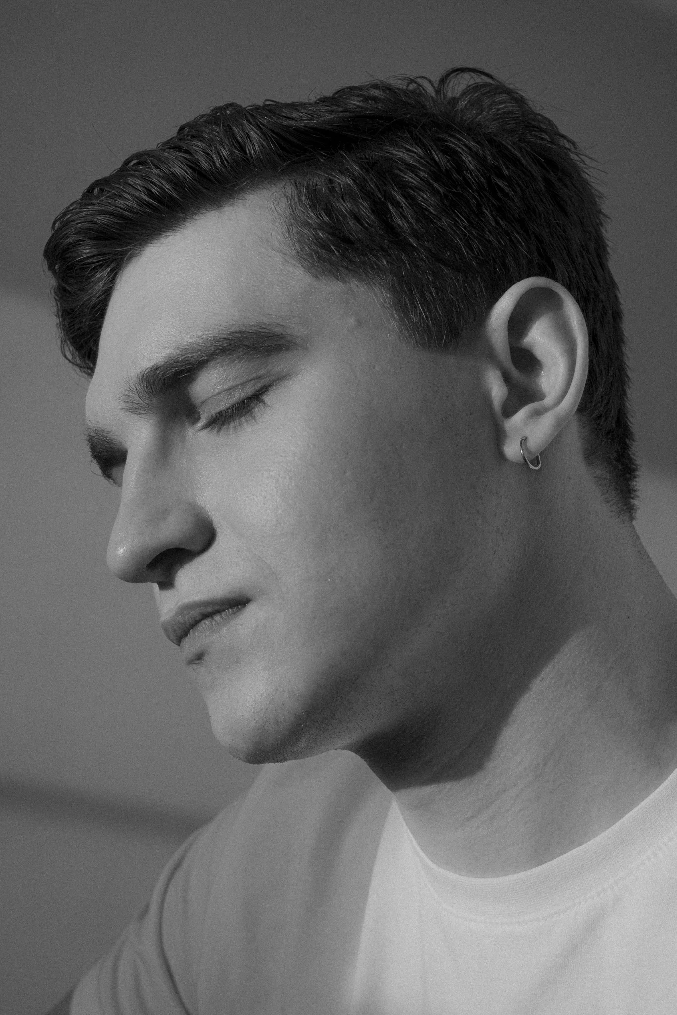 A black-and-white close-up photo of a young man with short, styled hair and a small hoop earring in his left ear, with his eyes closed and a calm expression.