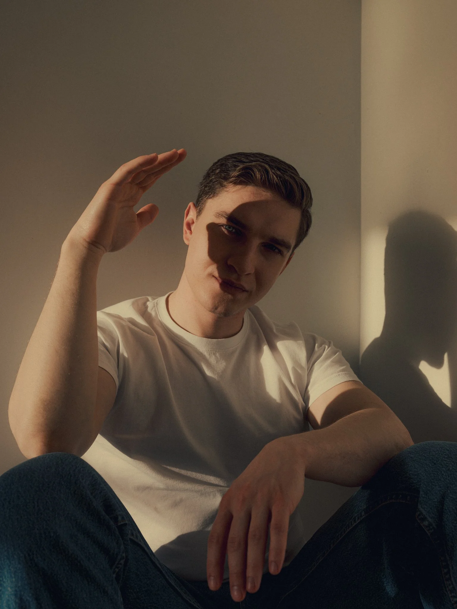 A young man with brown hair and wearing a white t-shirt, sitting against a wall with a shadow cast on it, raising his right hand near his head, with sunlight illuminating part of his face.