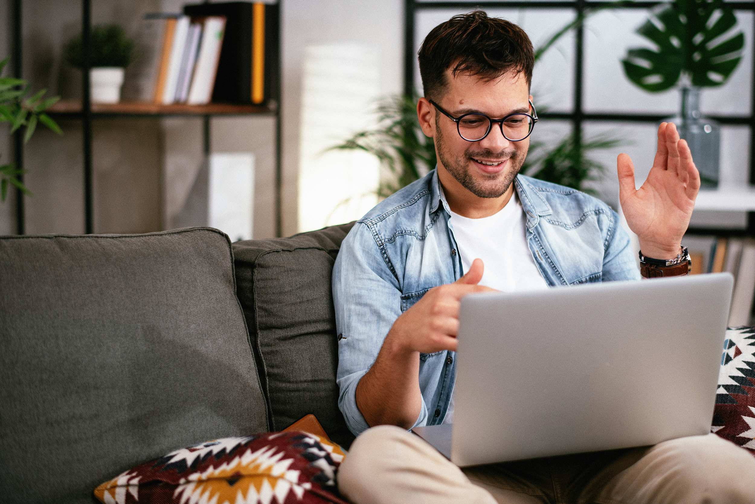 A young man with glasses and a denim shirt smiling and waving while sitting on a sofa with a laptop in front of him, in a home setting with plants and bookshelves in the background.