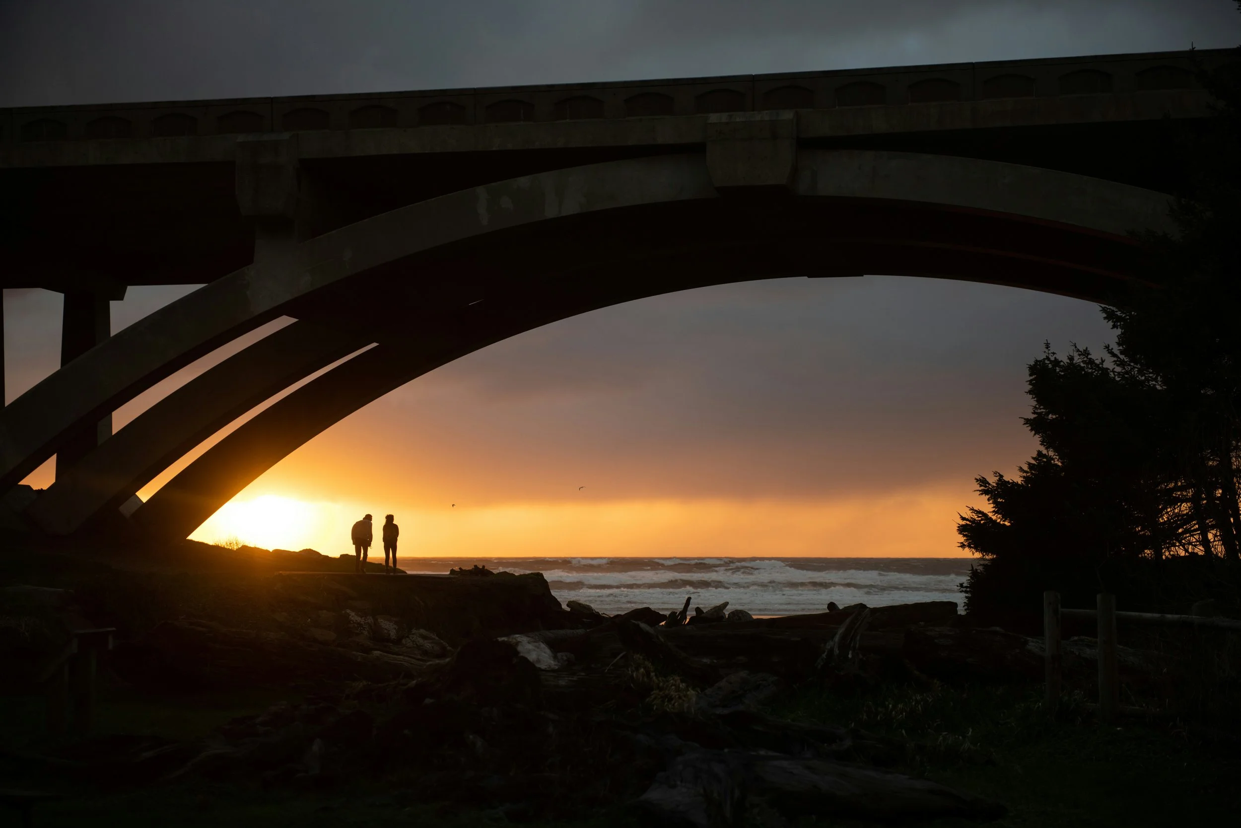Silhouettes of two people walking on a coastal area under a large bridge at sunset, with the sun setting over the ocean and a cloudy sky in the background.