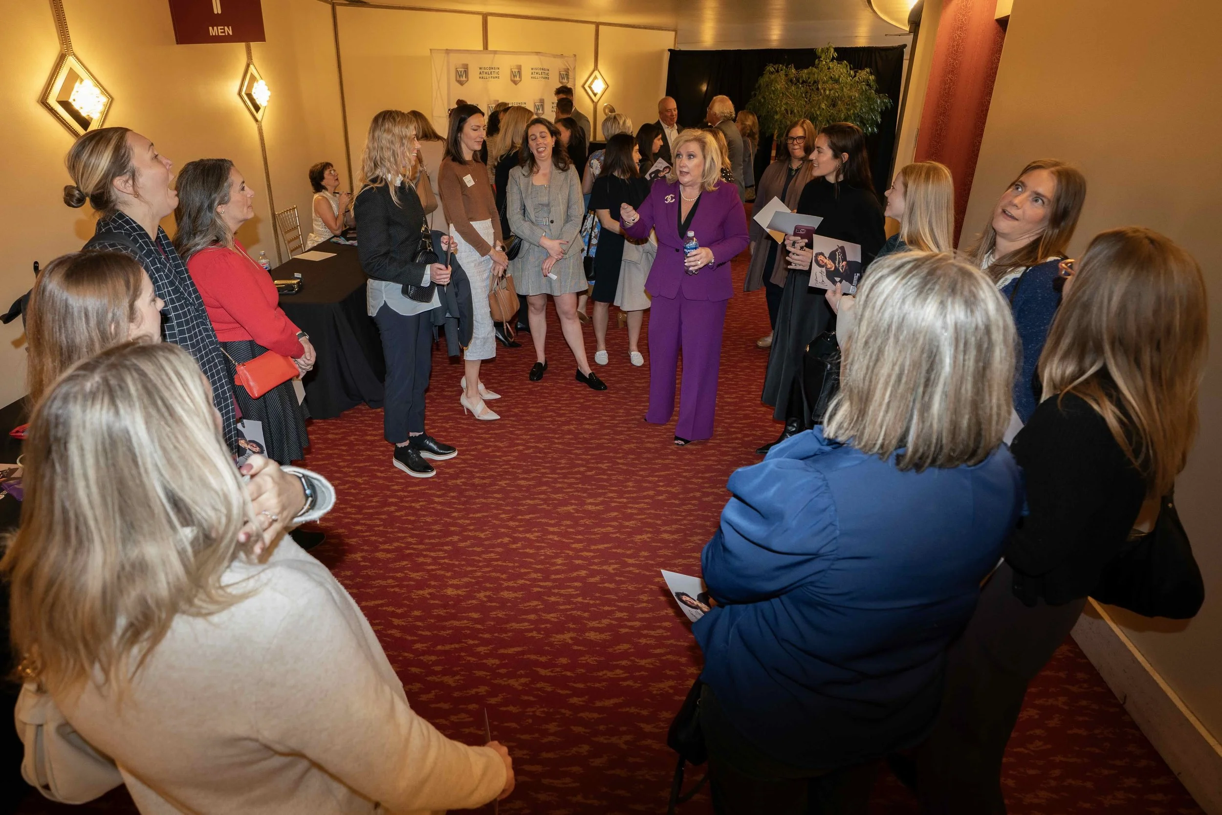 A woman in a purple suit is speaking to a group of women arranged in a circle. The setting appears to be a conference or networking event in a room with red carpet, warm lighting, and a banner in the background. The women are listening attentively an